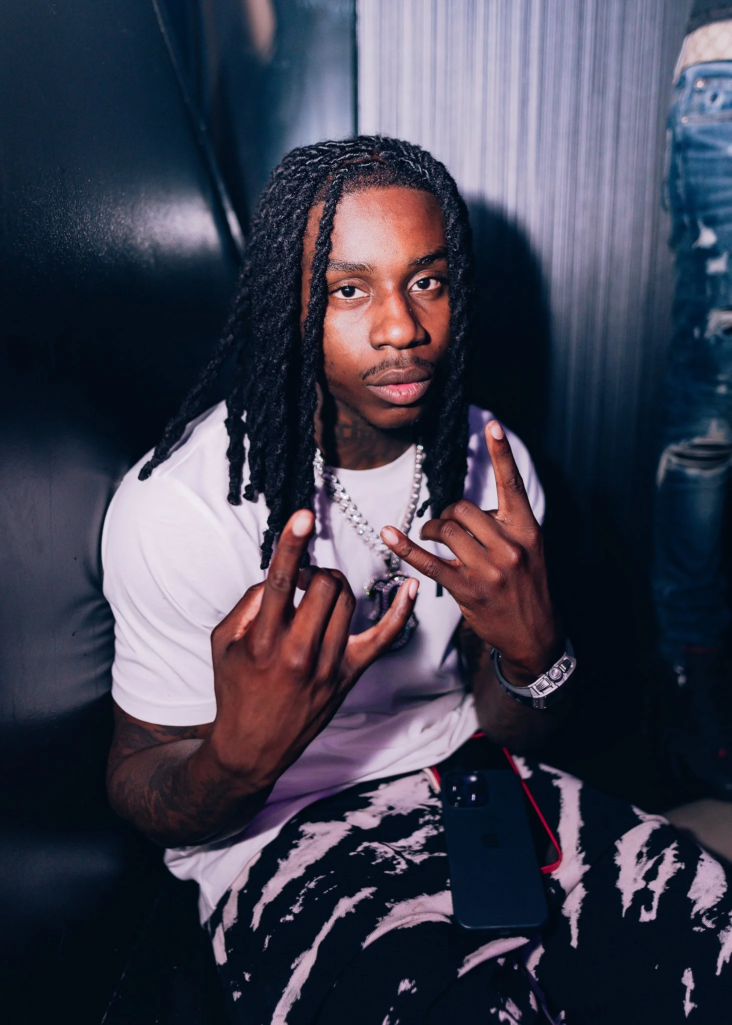 A young man with dreadlocks and tattoos is sitting on a dark couch, making hand signs with both hands. He's wearing a white t-shirt and patterned shorts, with jewelry and a watch. There is a water bottle on the right side.