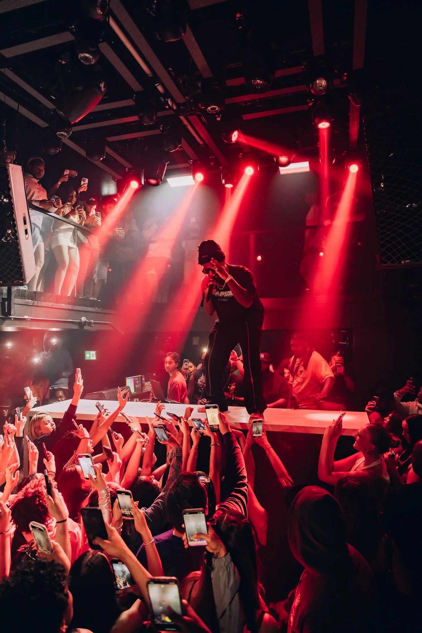 A performer on stage with red lights shining down at a concert, surrounded by an audience taking photos and videos.