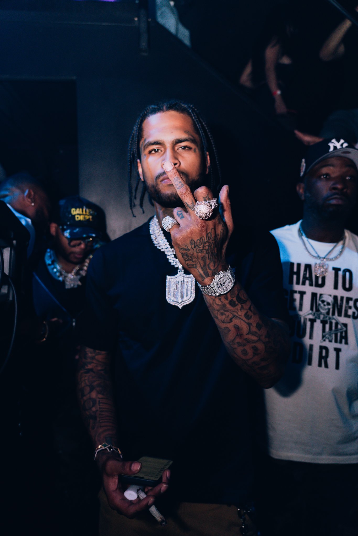 A man with braided hair and tattoos on his arms holding a cigarette with his middle finger raised. He is wearing large jewelry, including rings, a watch, and a chain. There are other people in the background, one wearing a cap and glasses, in a dimly lit environment.