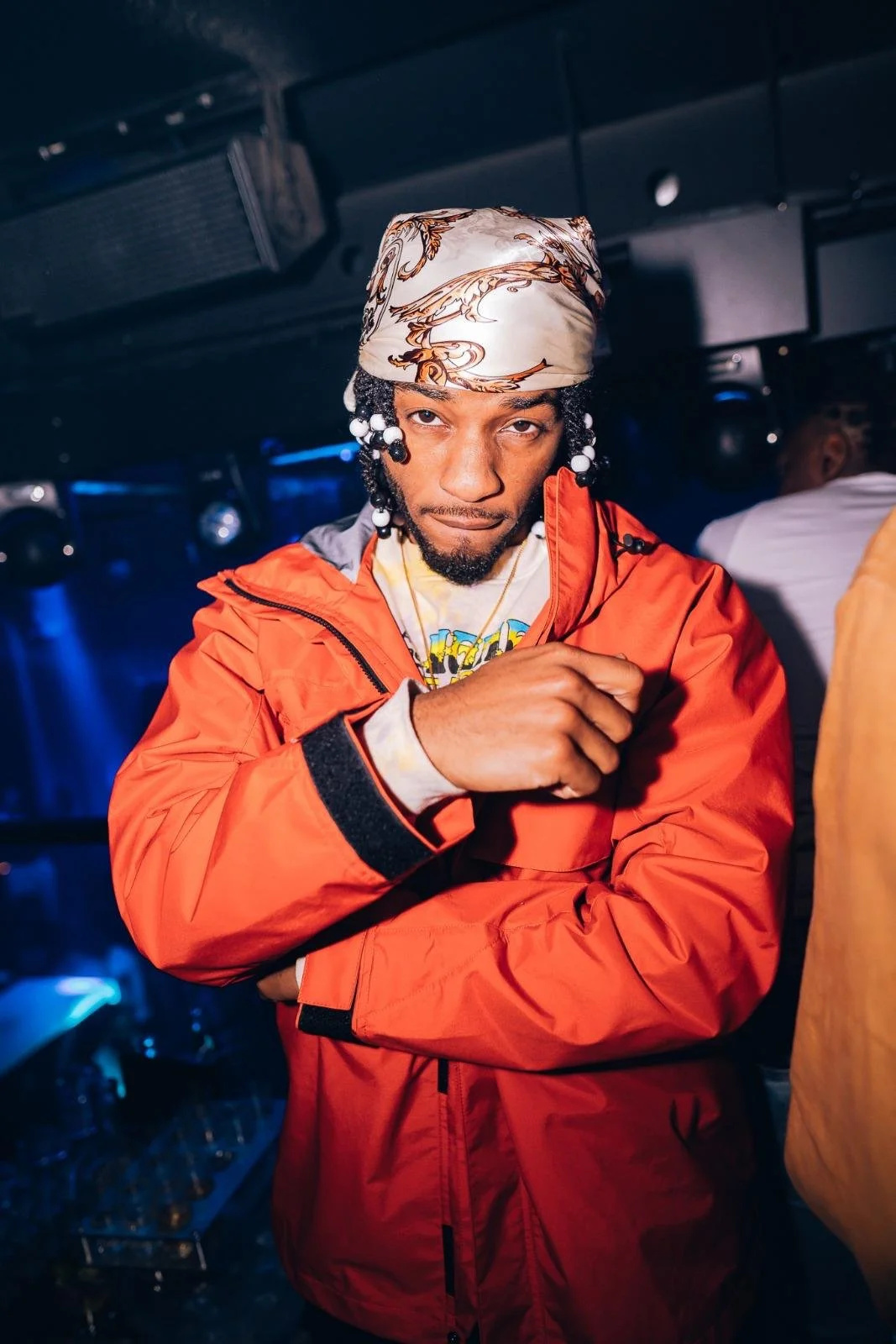 A man with a bandana on his head, wearing a red jacket, looking at the camera with a serious expression in a dimly lit indoor setting.