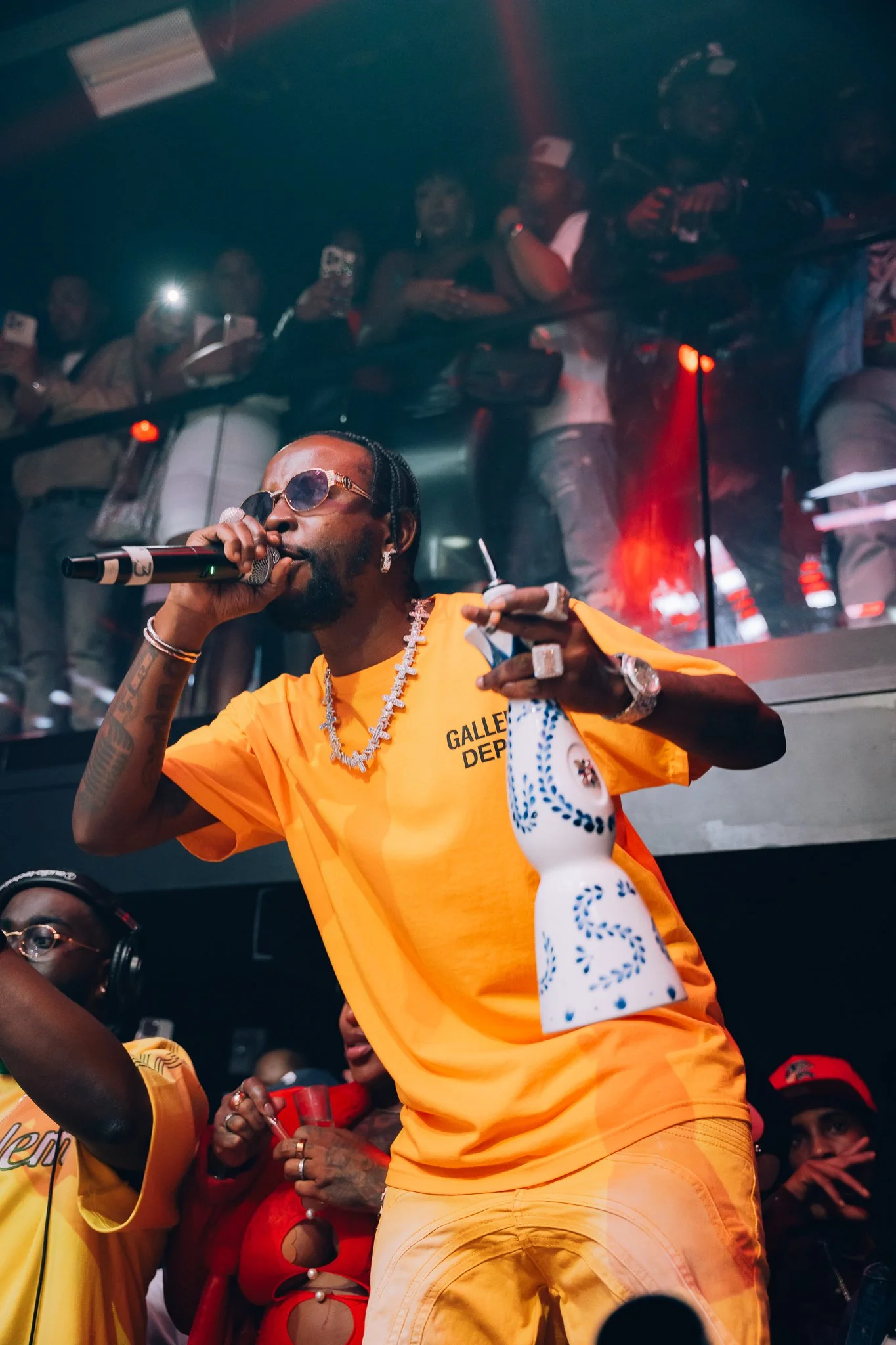 A man with sunglasses, earrings, and jewelry, dressed in an orange shirt, is holding a microphone and a white cloth, standing in front of a crowd at a lively event.