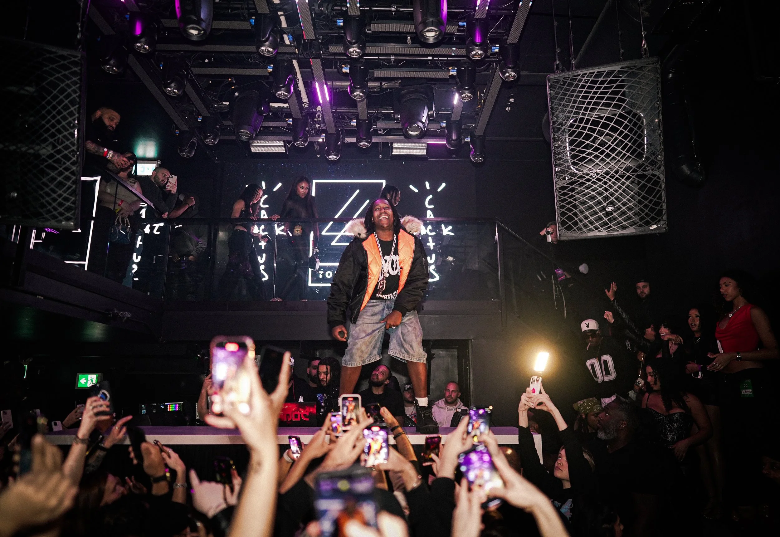 A performer on stage in a nightclub, posing and smiling, with a crowd of people taking photos and videos with their phones.