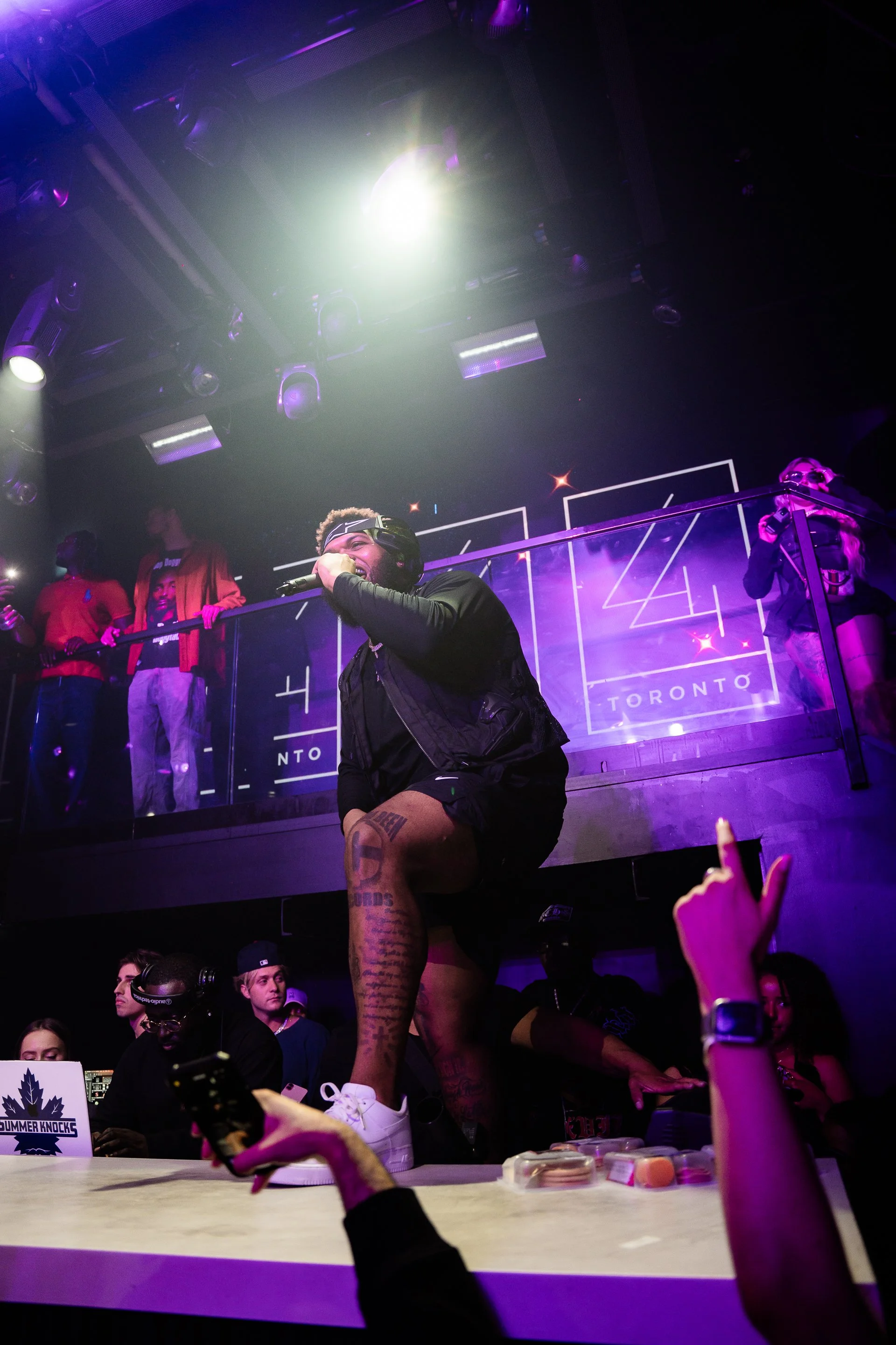 A lively scene at a nightclub or concert featuring a male performer with a microphone, standing in front of a purple-lit stage with other people in the background. The stage has a sign that reads 'Toronto' and 'Summer Knocks'. Audience members are visible, some recording with their phones.