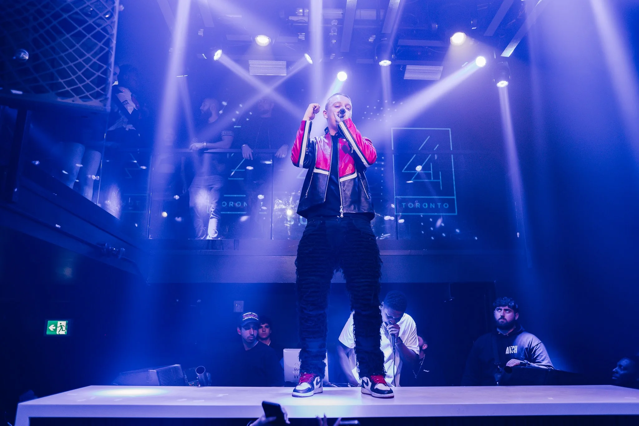 A performer, a young person, standing on a stage with a microphone, wearing a red and black jacket and sneakers, with spotlights shining down on them. There are people in the background behind a glass barrier, and stage equipment and an exit sign are visible.