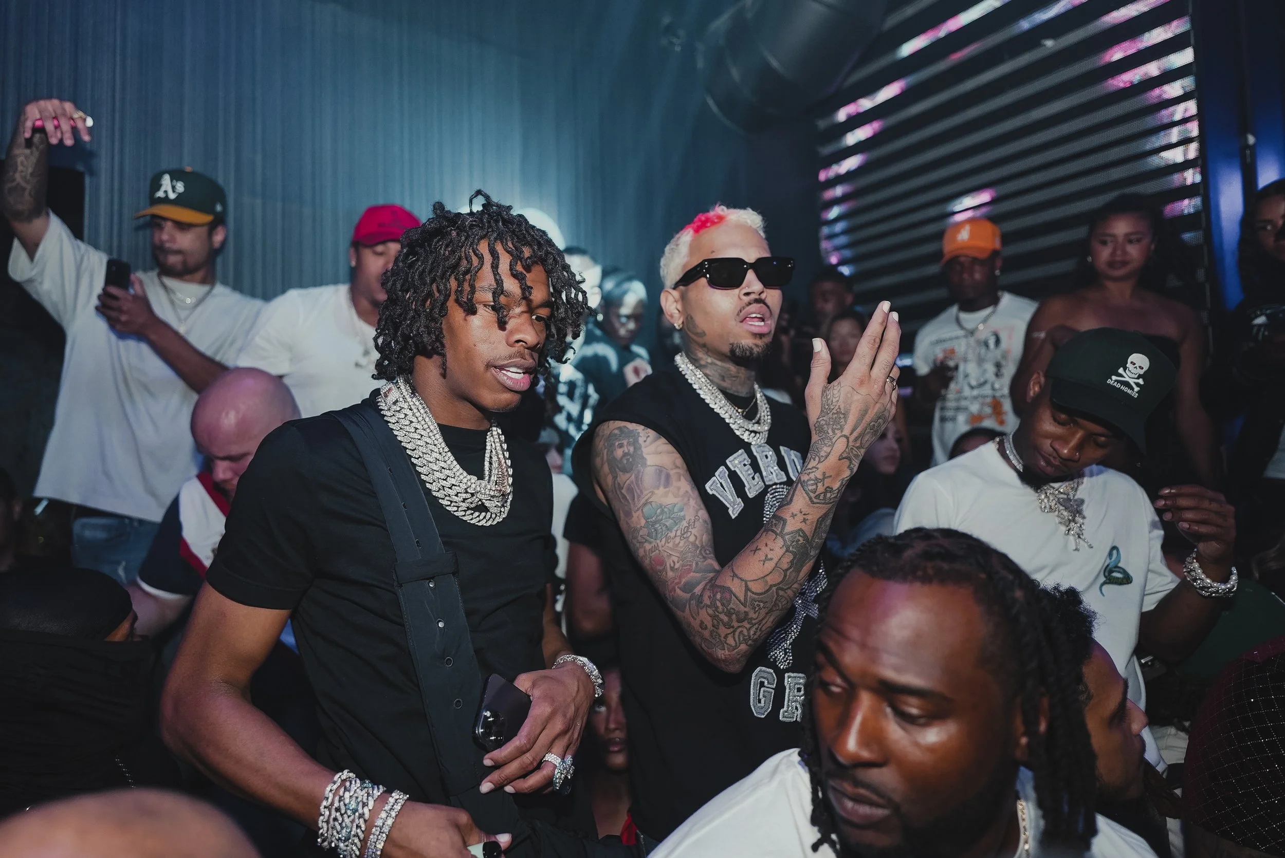 Two men with jewelry and tattoos attending a music event, surrounded by other attendees in a dim, colorful venue.
