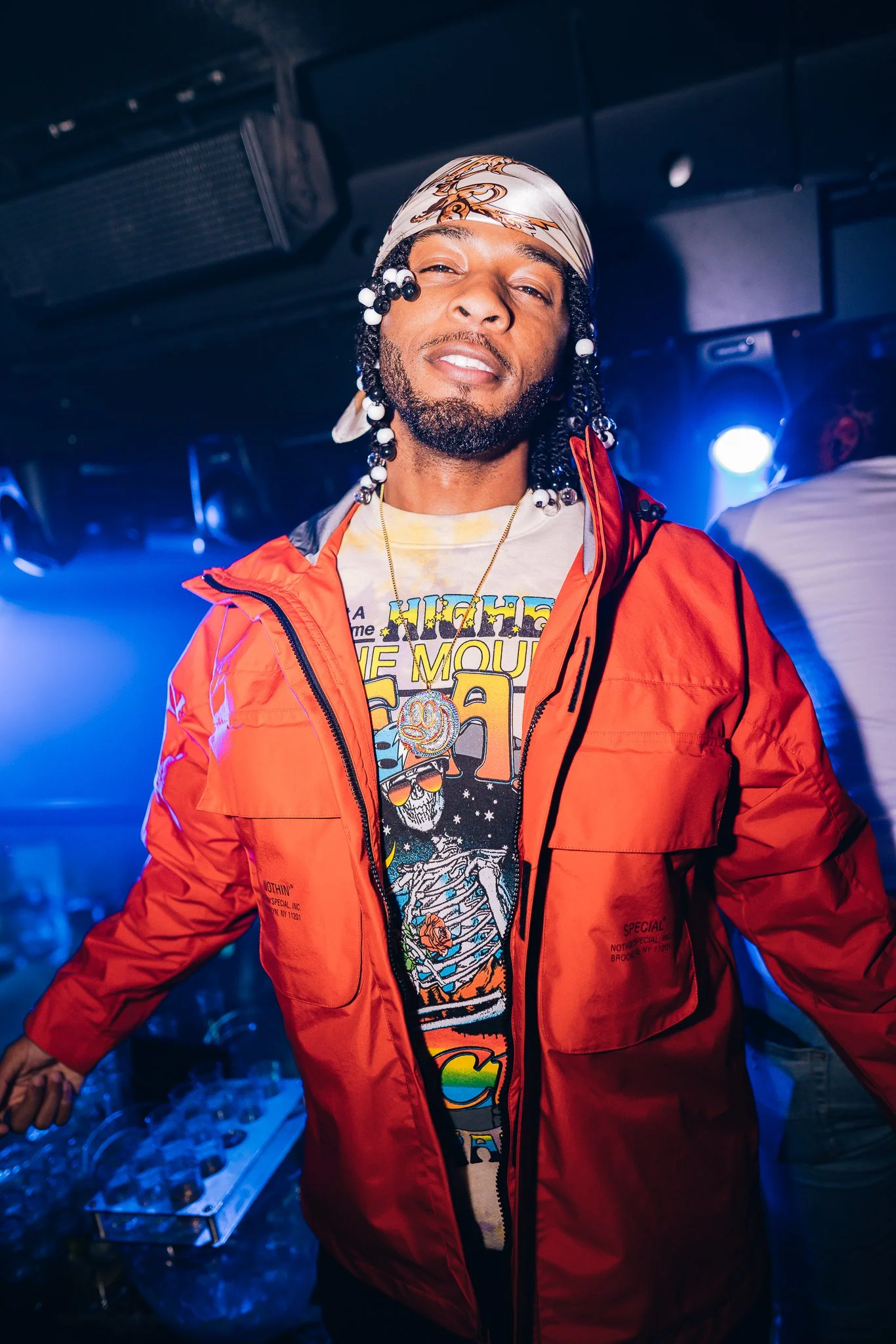 A man with medium skin tone, black beaded dreadlocks, and a light-colored bandana with a design, wearing a vibrant orange jacket over a graphic T-shirt, stands in a dimly lit room with blue lighting, smiling slightly at the camera.