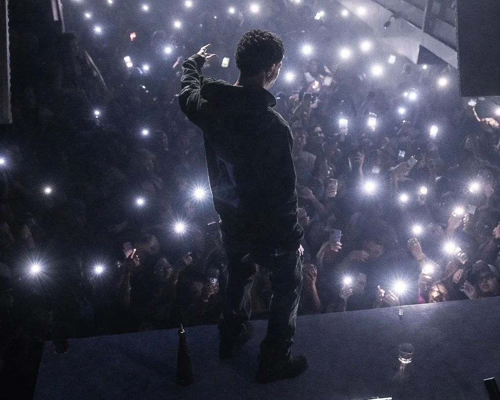 A person stands on stage with their back to the camera, facing a large crowd holding up smartphones with flashlights on, in a dimly lit venue.