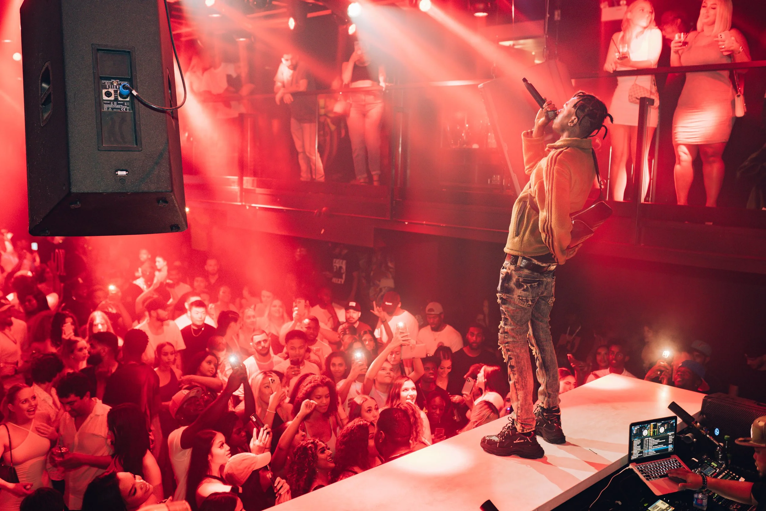 A live performer on stage with a microphone, wearing a tan hoodie and ripped jeans, facing a large crowd in a club or concert venue with red lighting. Several women are watching from a balcony above, and a DJ setup is visible at the front of the stage.