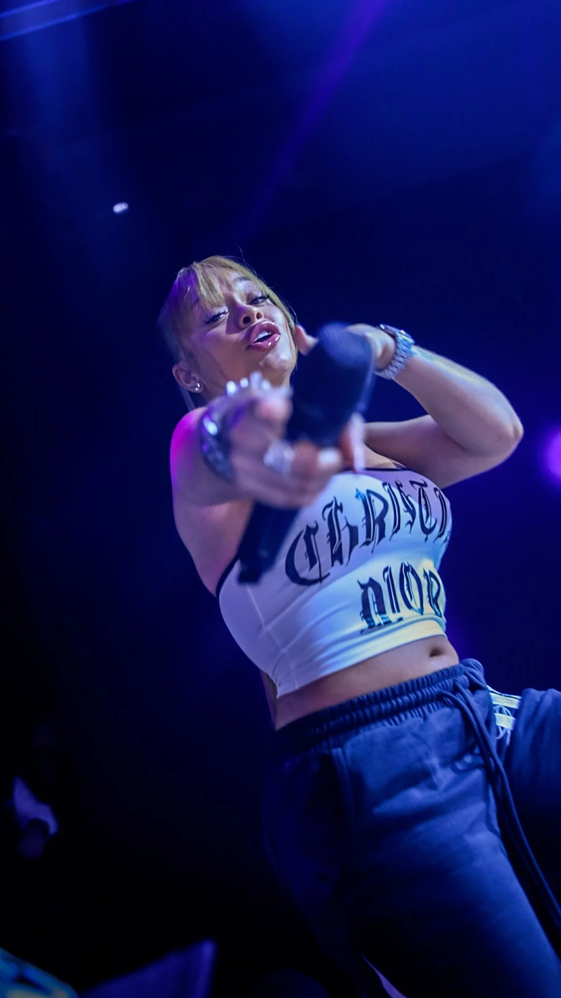 A woman on stage holding a microphone, wearing a crop top and pants, under purple and blue stage lighting.