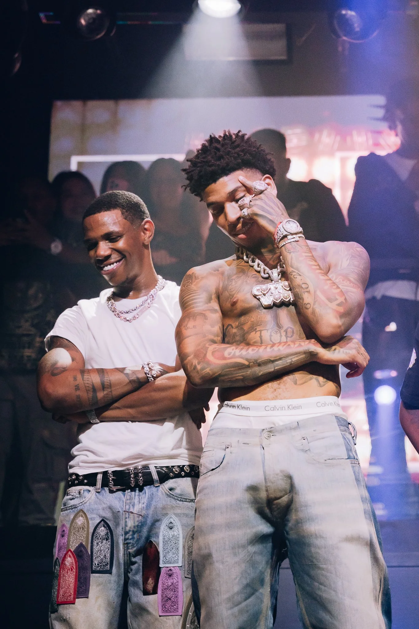 Two young men are on a stage with one shirtless, showing tattoos, wearing light jeans and jewelry, striking a pose next to another man in a white t-shirt and jeans with jewelry. The background suggests a club or concert setting.