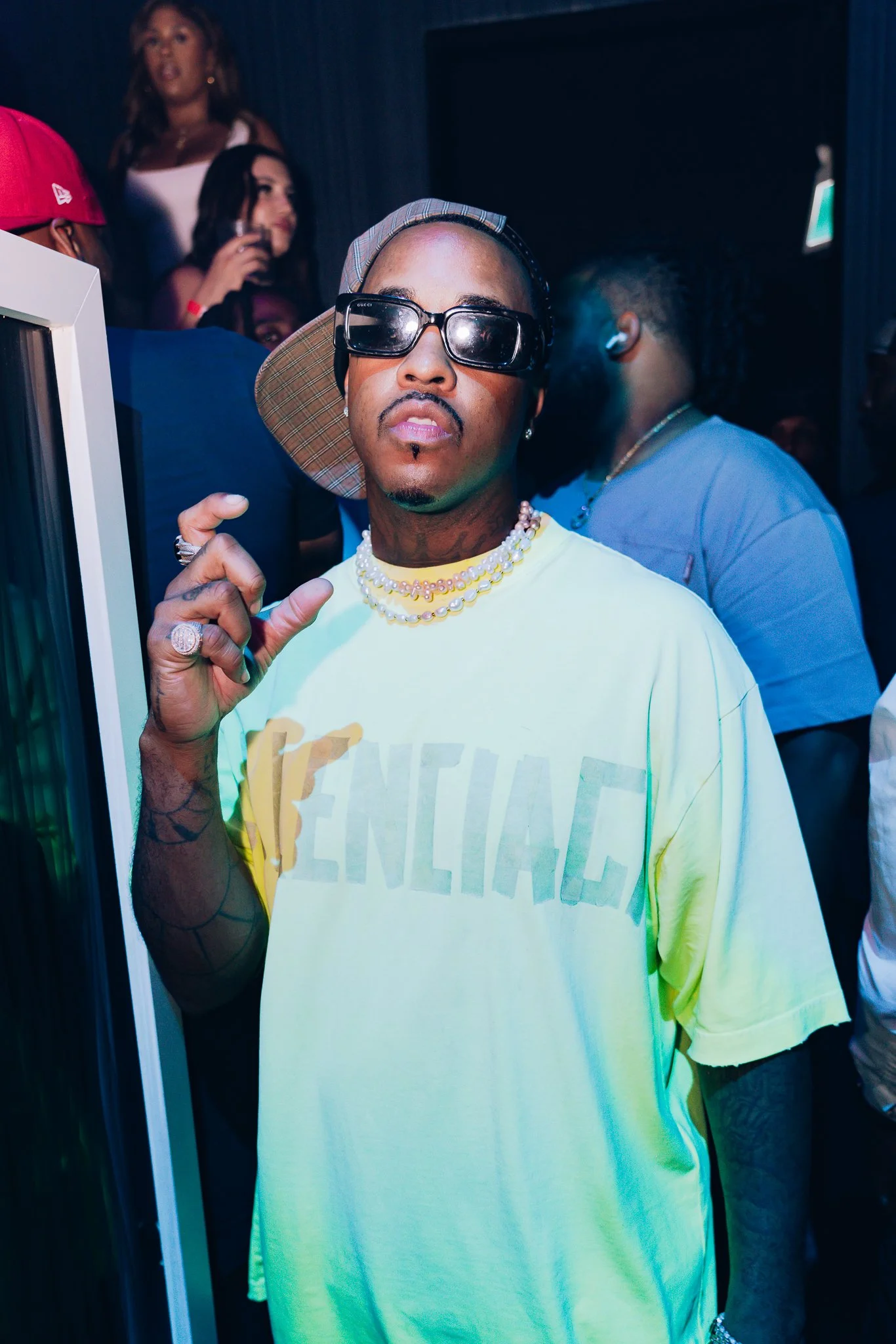 A man wearing large sunglasses, a colorful cap, and layered jewelry is pointing at the camera. He is in a room with other people in the background.