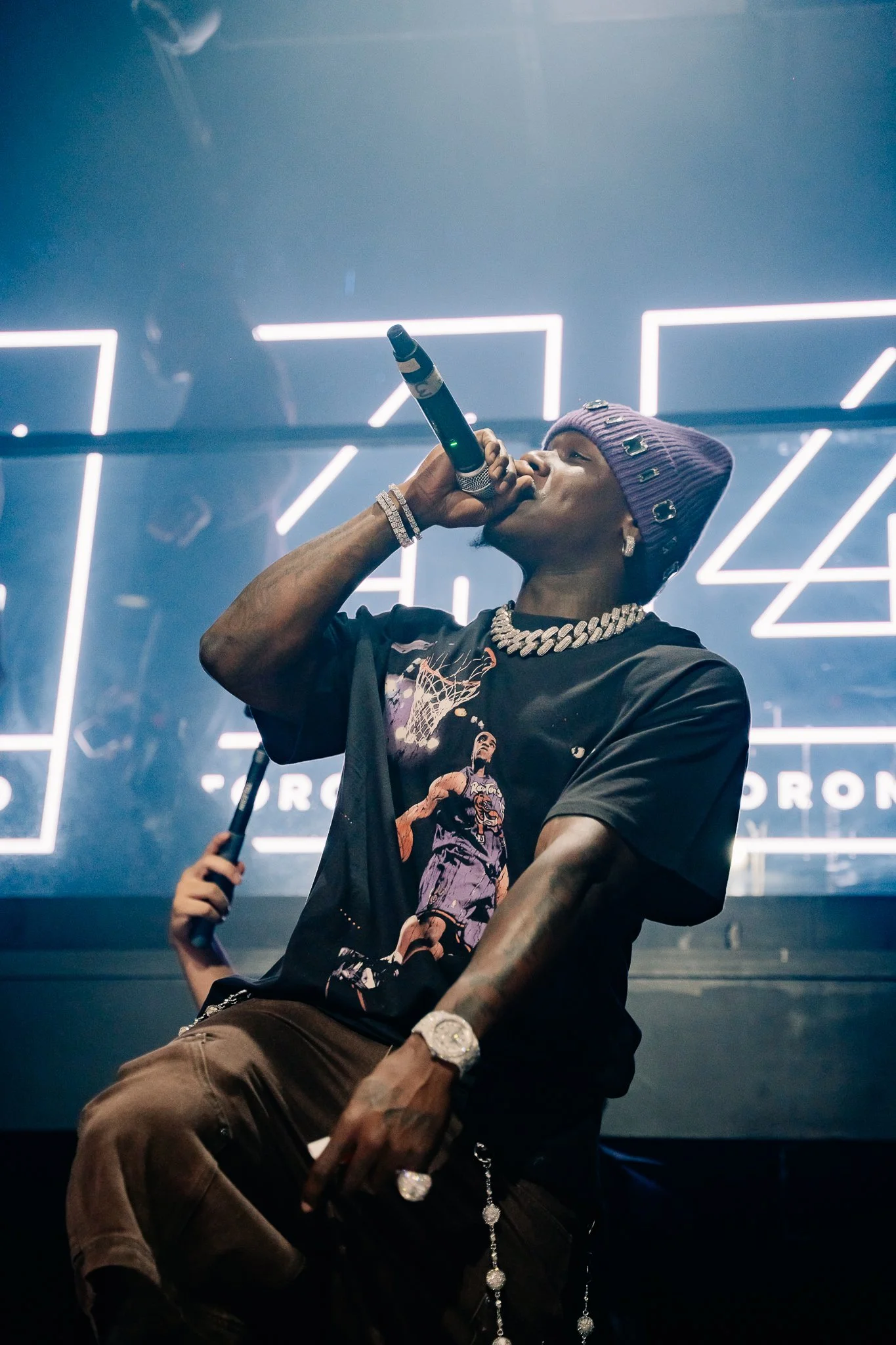 Performer with a purple beanie, diamond earrings, chains, and a graphic T-shirt, singing into a microphone on stage with neon lights in the background.