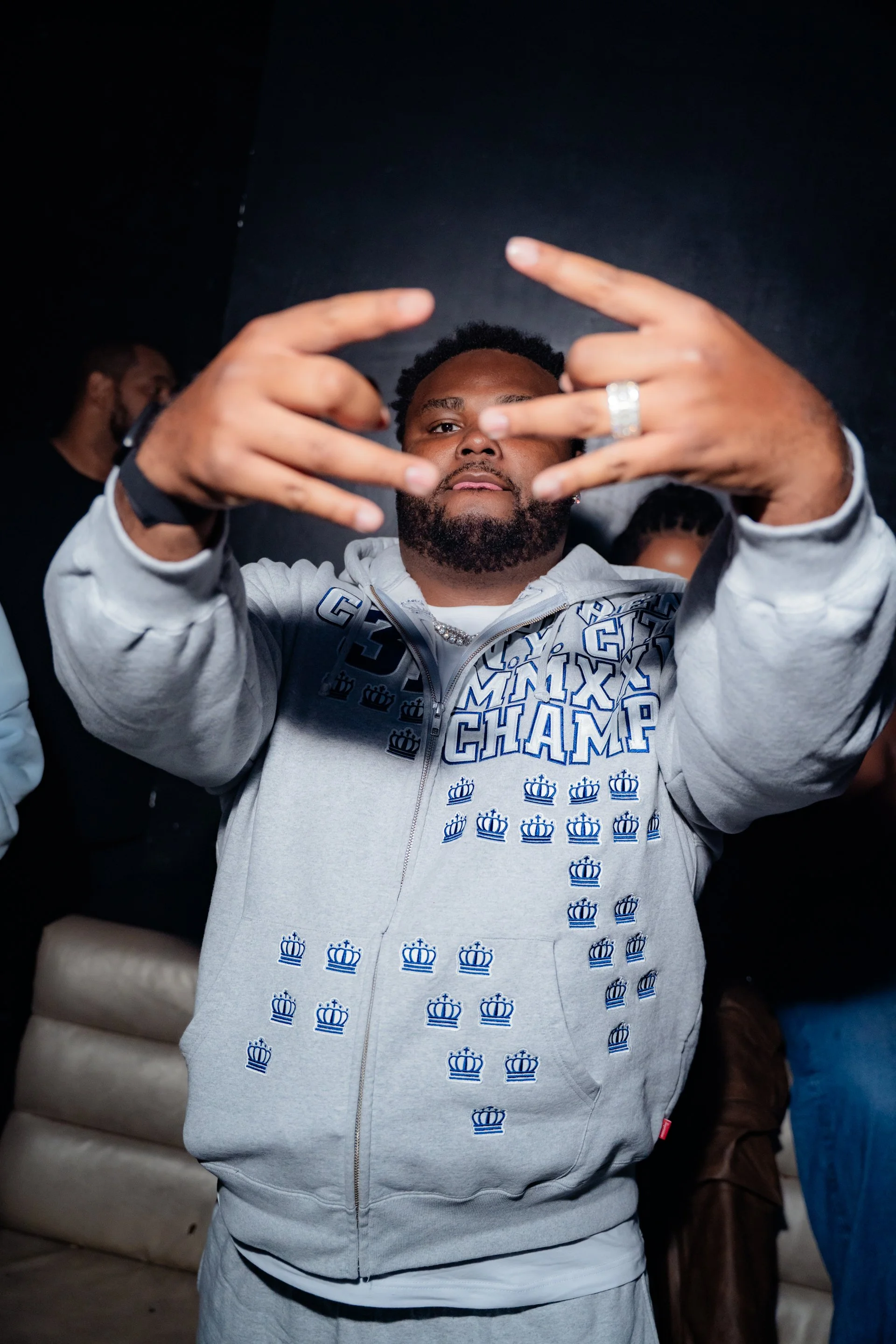 A man with a beard making a gesture with his hands in front of the camera. He is wearing a light gray hoodie with a pattern of blue crowns and the words 'King's Champ'. There are other people partially visible in the background.