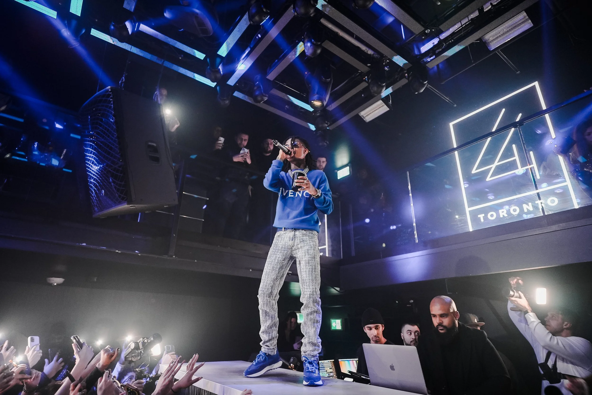 A male performer standing on a stage, wearing patterned pants, a blue sweatshirt with the word 'GIVENCHY,' and holding a microphone. The stage is equipped with professional lighting, and there are numerous people in the audience taking photos and videos. The background features a large display with a sign that says 'TORONTO' and the number '44.'
