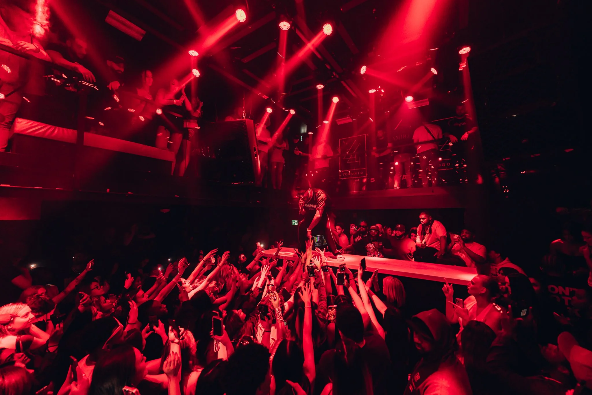 A DJ mixes music on a turntable on stage while a crowd dances and raises their hands at a nightclub with red lighting.