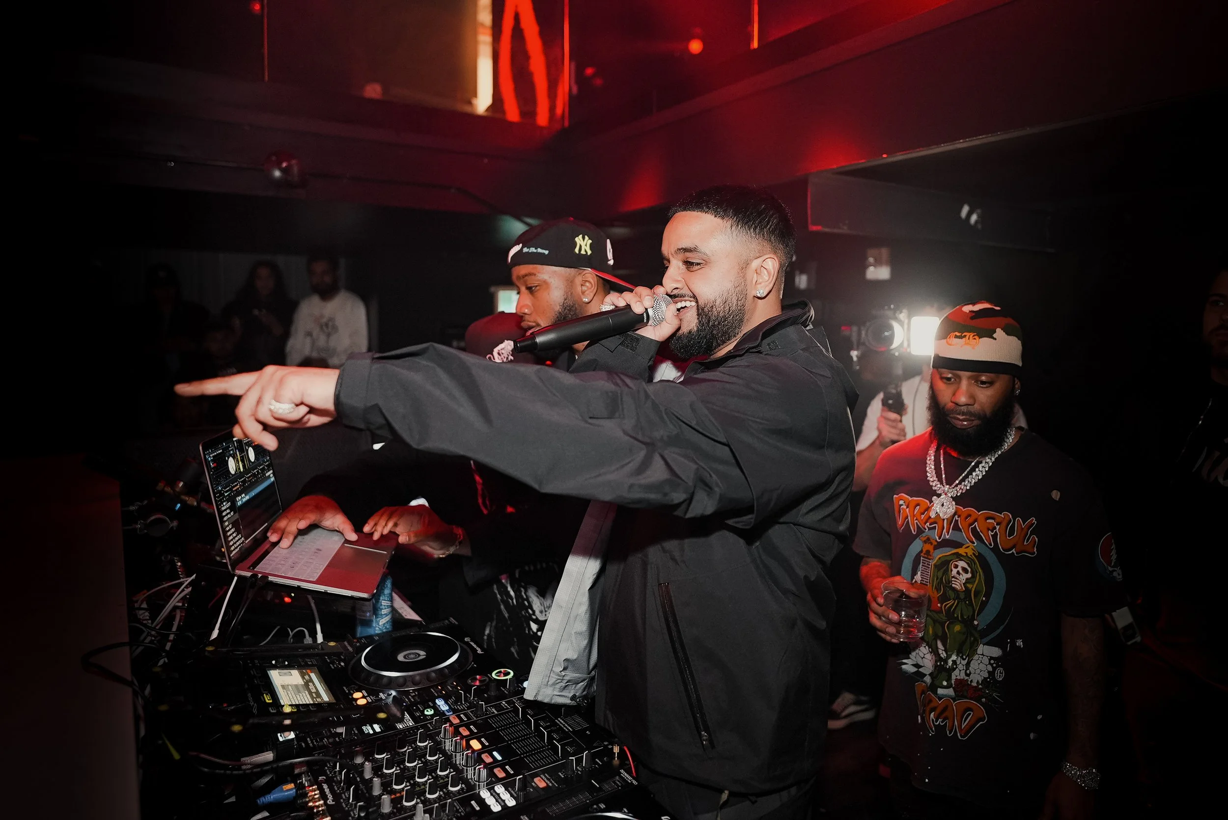 A DJ with a microphone in hand performing at a nightclub with red lighting, surrounded by other people, including one wearing a colorful graphic T-shirt and a beanie, and a DJ setup with turntables and a laptop.