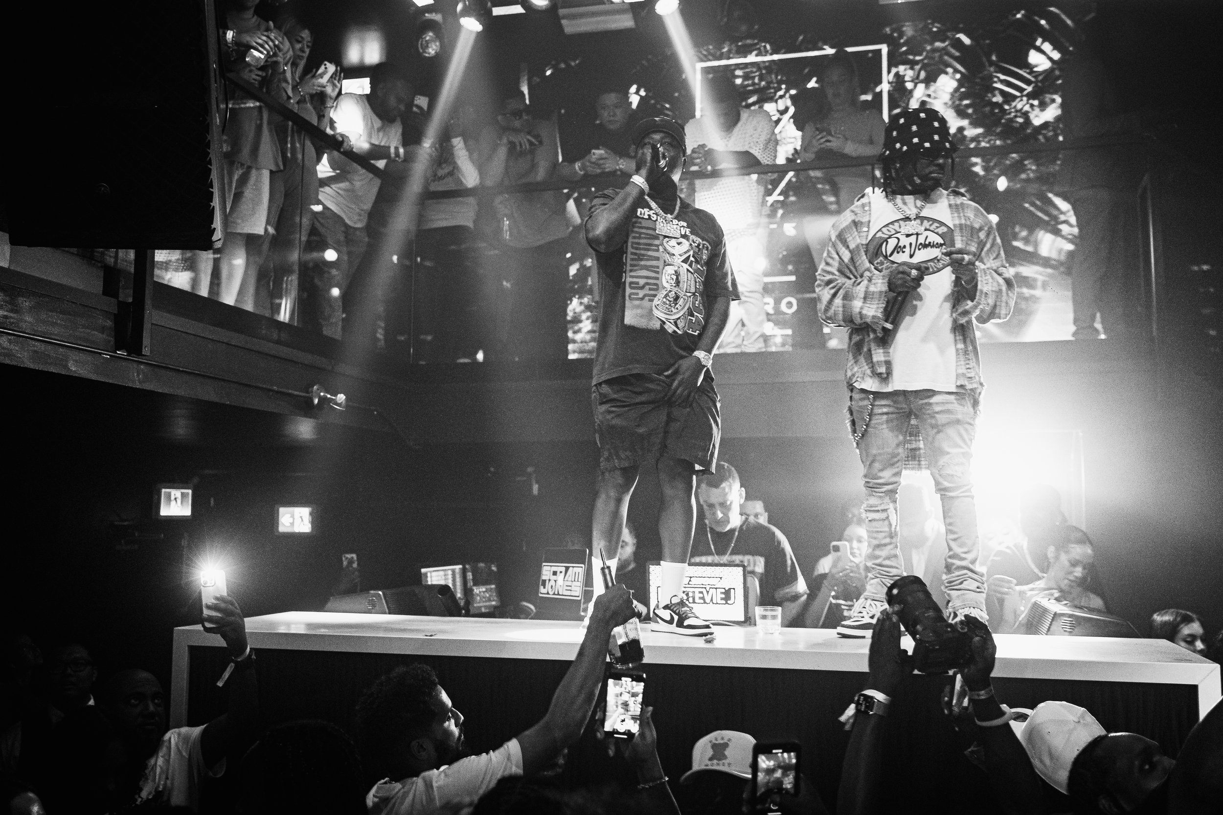 A black-and-white photograph of a nightclub or concert setting featuring two male performers on stage with audience members watching and holding up smartphones. One performer is wearing a cap and patterned shirt, the other in graphic clothing. Bright stage lights illuminate the scene.