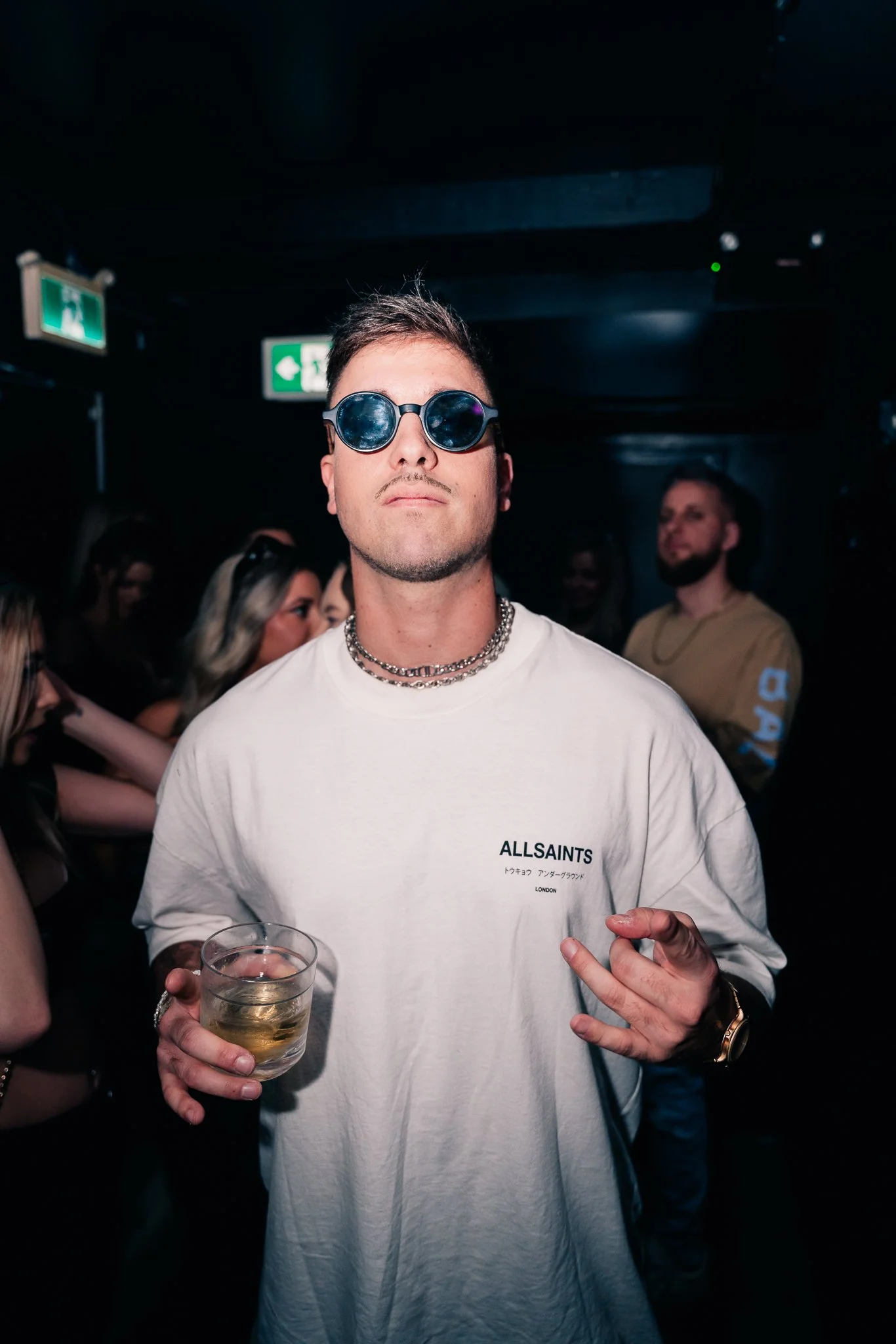 A young man wearing round sunglasses, a white sweatshirt with 'ALLSAINTS' printed on it, a chain necklace, holding a glass of beer, and making a peace sign with his hand at a social event in a dimly lit space.