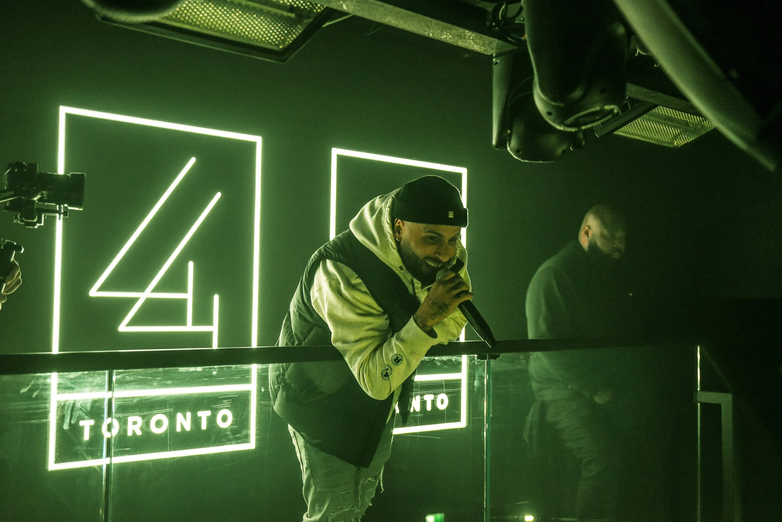 A person with tattoos on their hand is smiling and holding a microphone while standing in front of illuminated signs that say 'TORONTO' and 'TRONTO' in a dark room with green lighting.