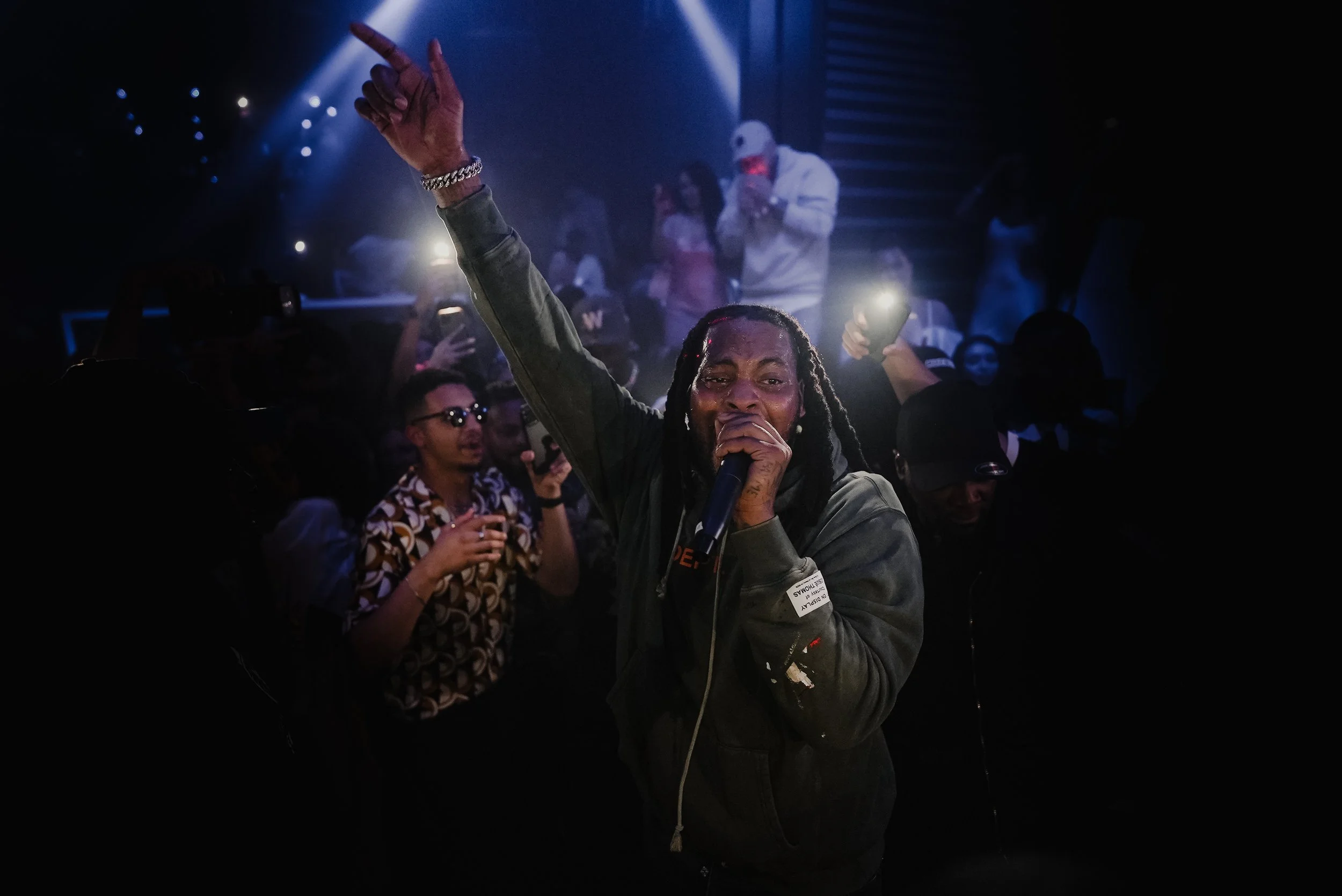 A performer singing into a microphone on stage with a lively crowd in the background, some attendees are taking photos or videos, and blue stage lights add to the lively atmosphere.