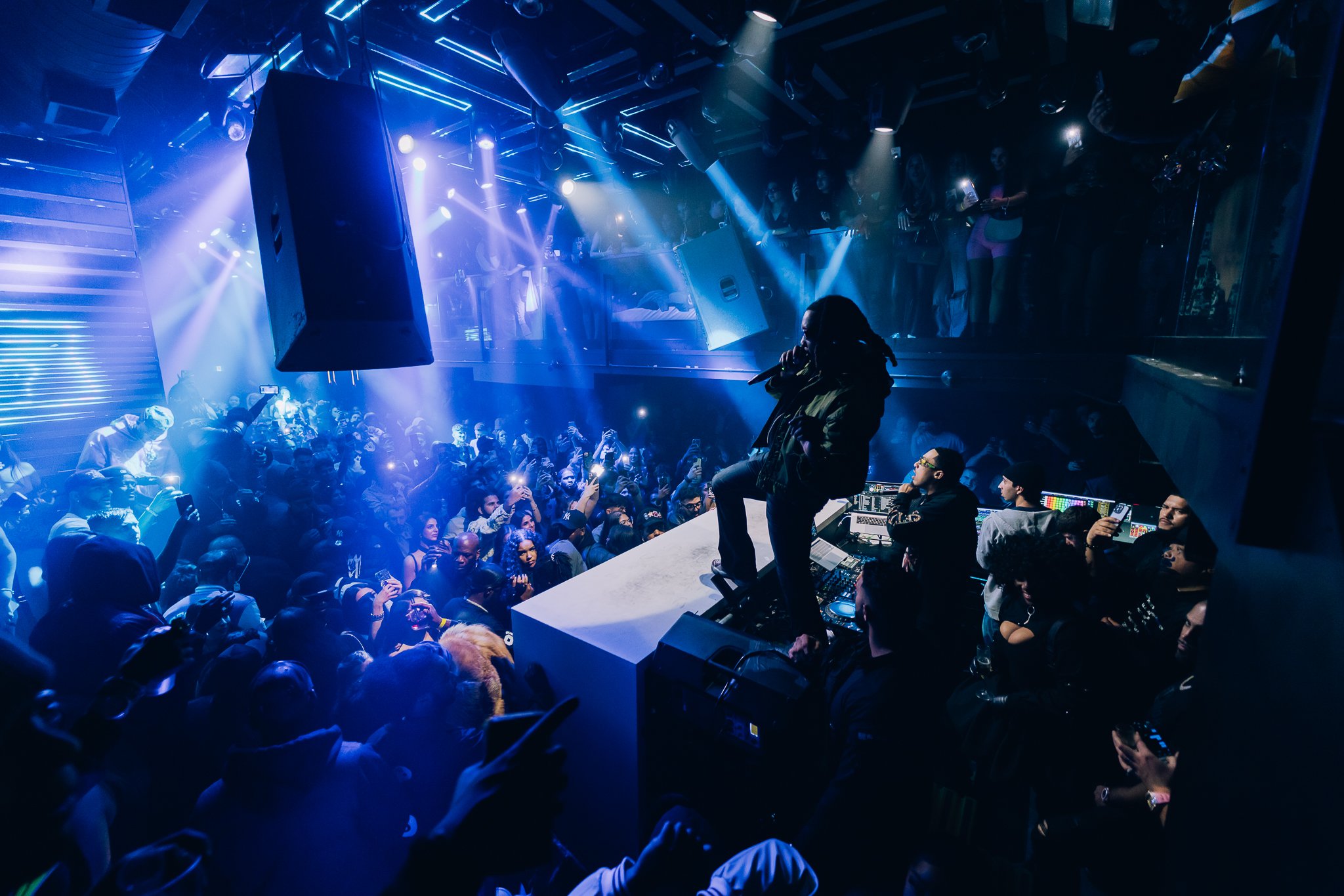 A crowded DJ nightclub scene with a performer on stage, surrounded by blue and purple stage lights, and a lively audience taking photos.