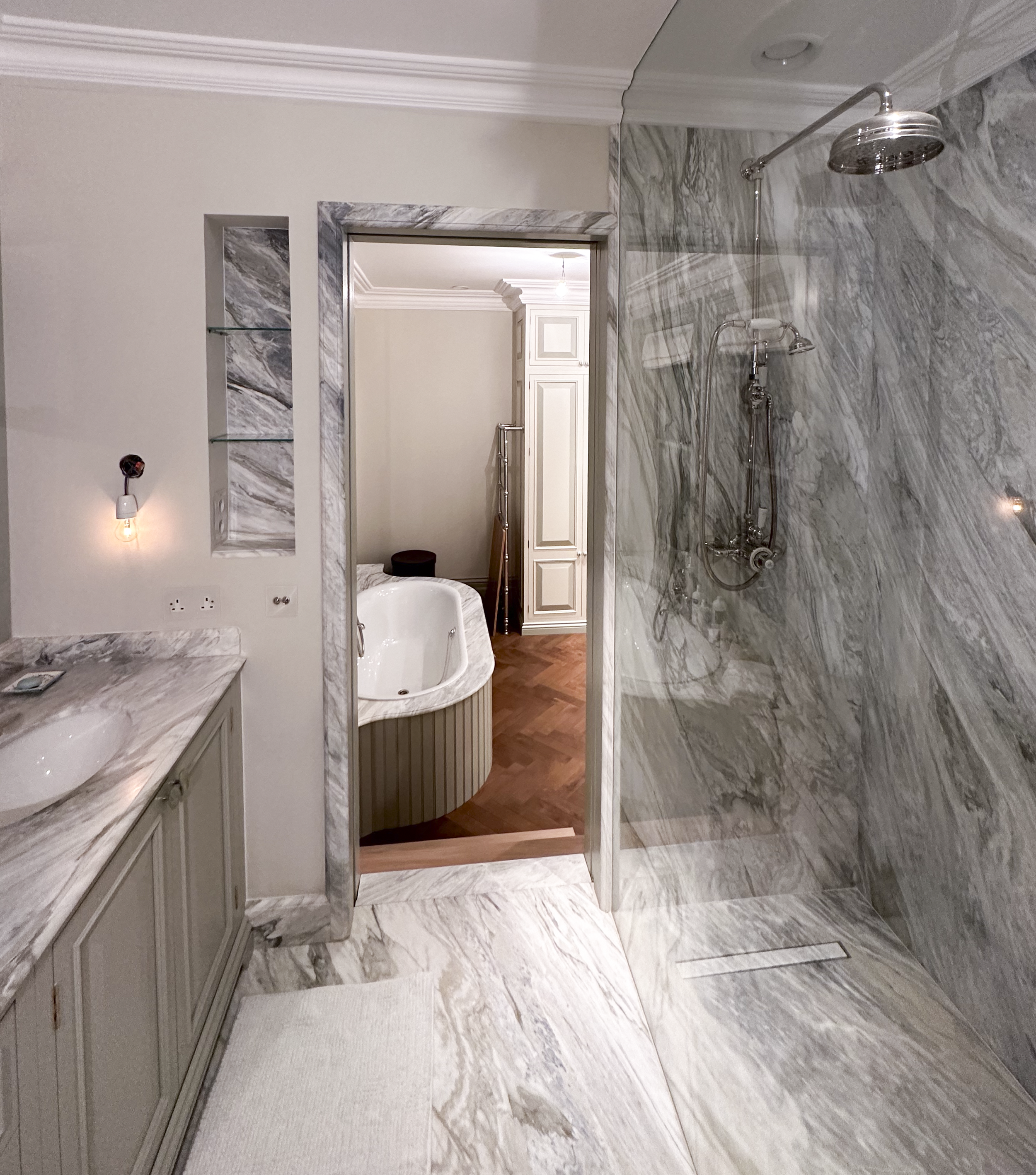 A luxurious bathroom with marble walls and floor, featuring a walk-in shower with a rain showerhead, a vanity with a marble countertop, and a doorway leading to a room with a bathtub and wooden floors.