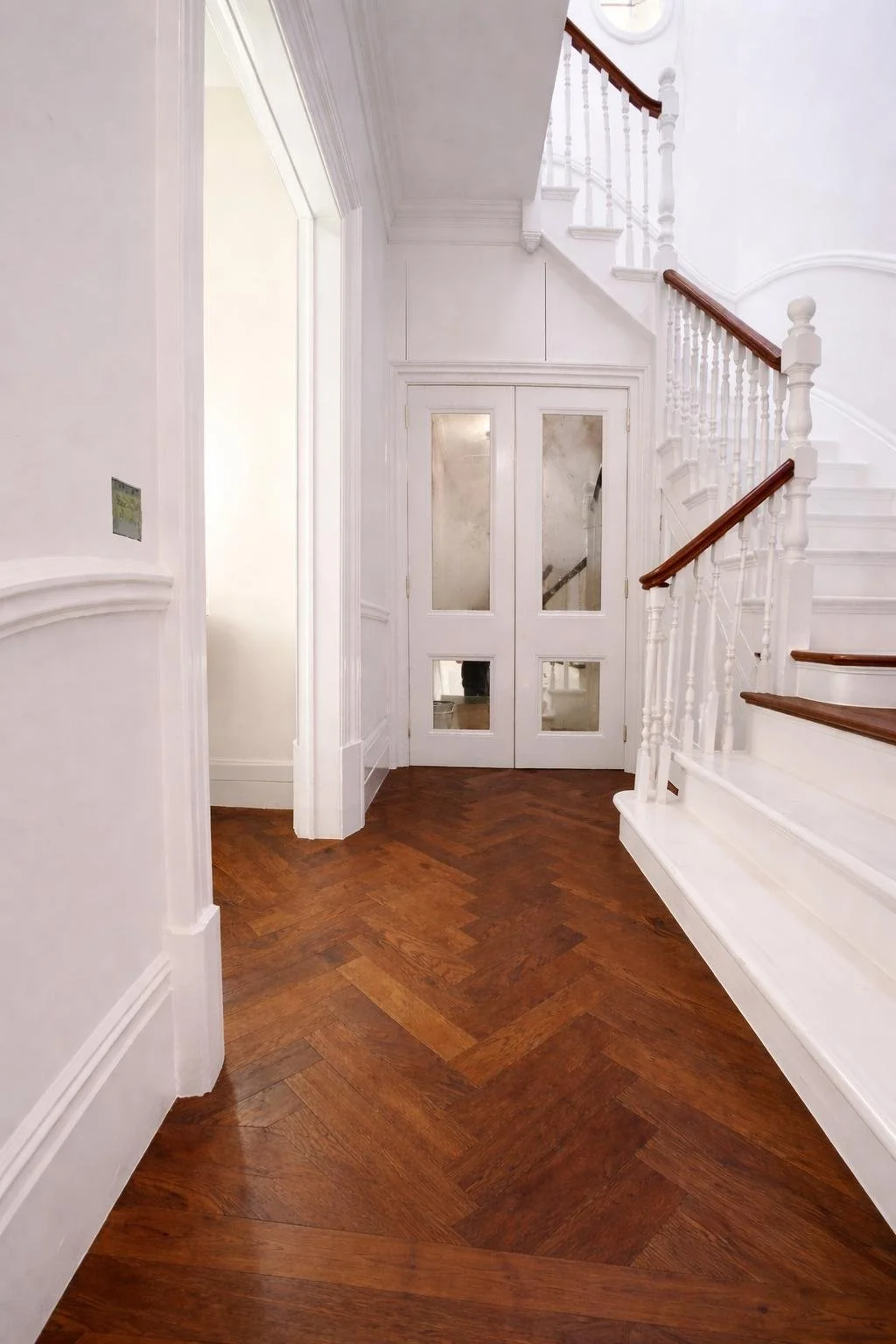 Entryway of a house with white walls, wooden floors, a staircase with white balusters and wooden handrails, and a tall double door with glass panels.