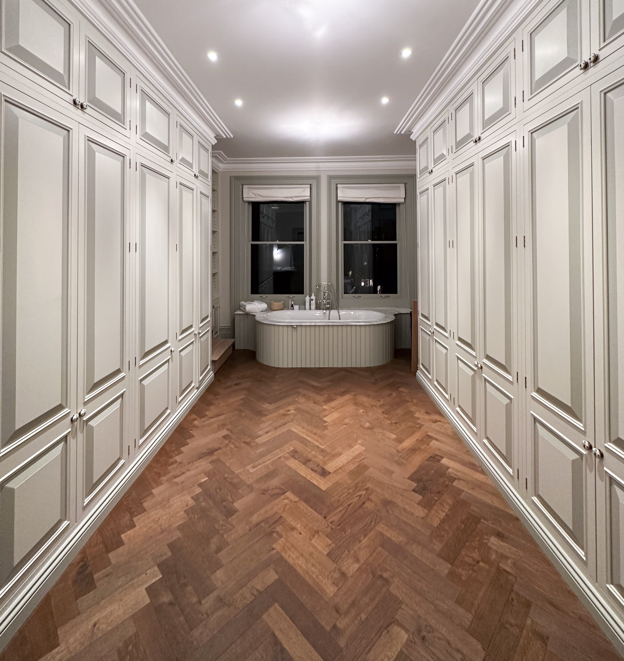 A spacious bathroom with built-in beige cabinets on both sides, a wooden herringbone floor, and a bathtub near the window at the back.