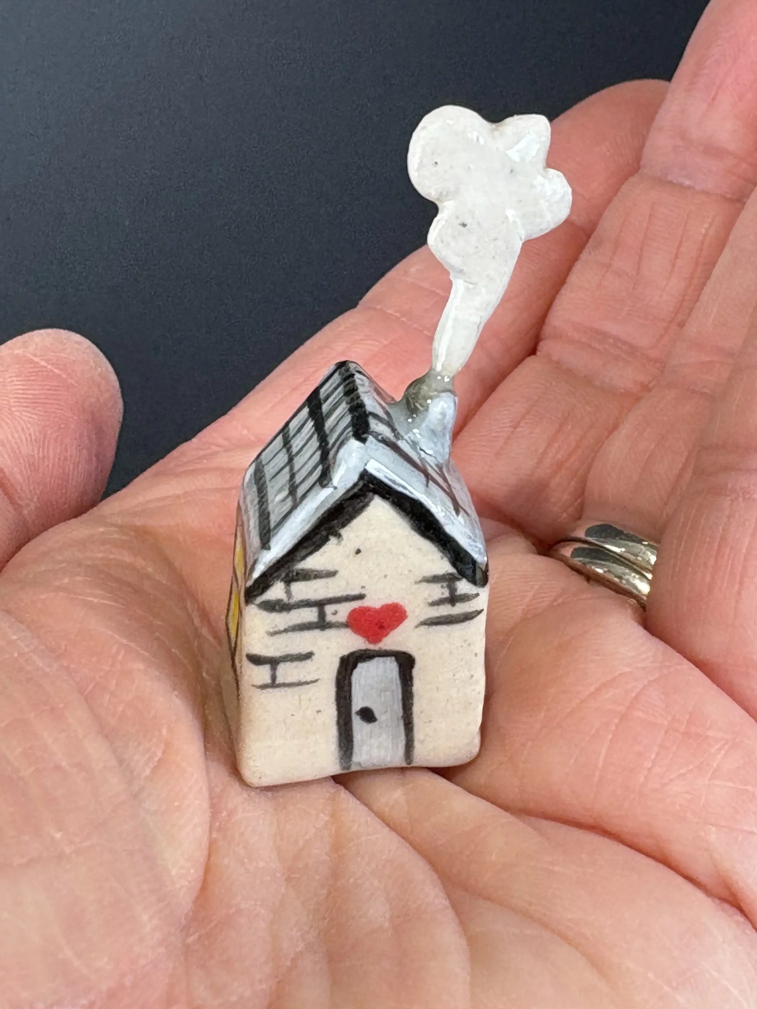 Small ceramic house sculpture with chimney smoke coming from a painted black roof and a red heart on the front, held in the palm of a person's hand.