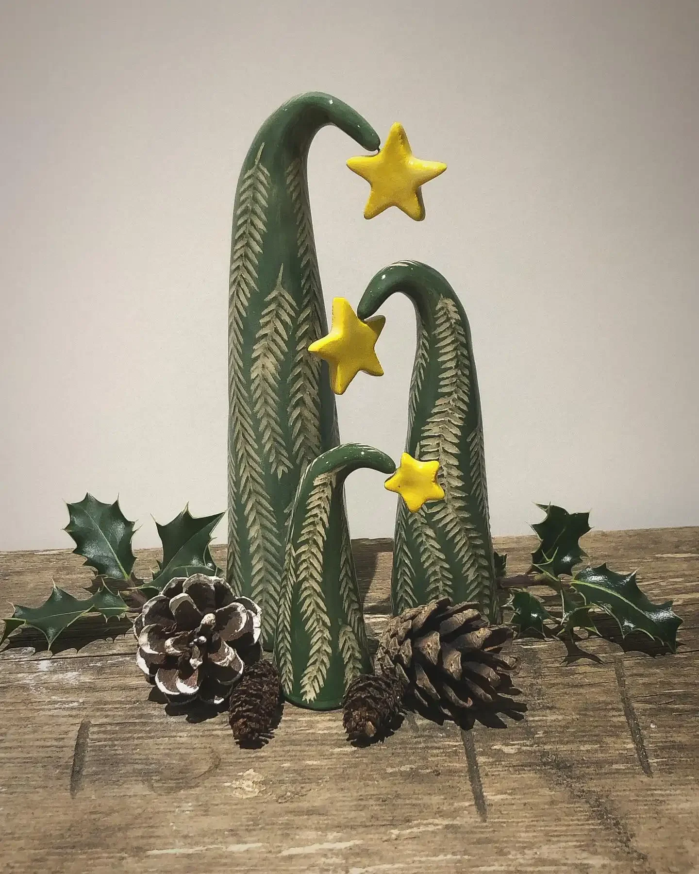 Decorative Christmas centerpiece featuring three green ceramic trees with yellow star toppers, surrounded by pinecones and holly leaves on a wooden surface.