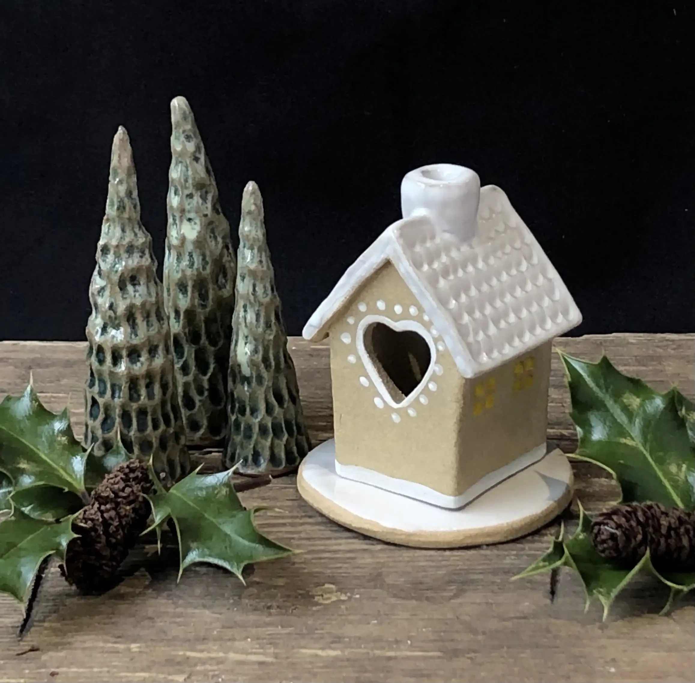 Christmas ceramic house ornament with cutout white heart window, three green textured cone-shaped trees, holly leaves, and pine cones on a wooden surface against a black background.