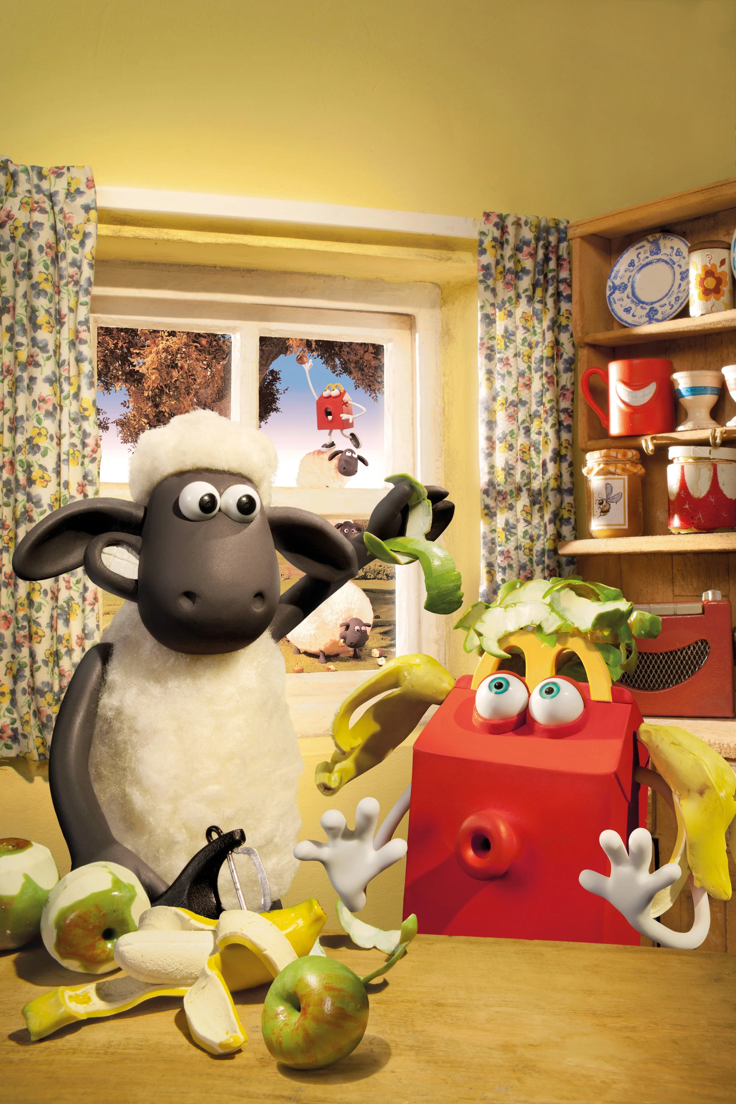 Aardman animated Shaun the Sheep and the Happy Meal box character in a kitchen. The table is covered with peeled bananas and apples. The background shows a window with curtains and a view of animated sheep outside.