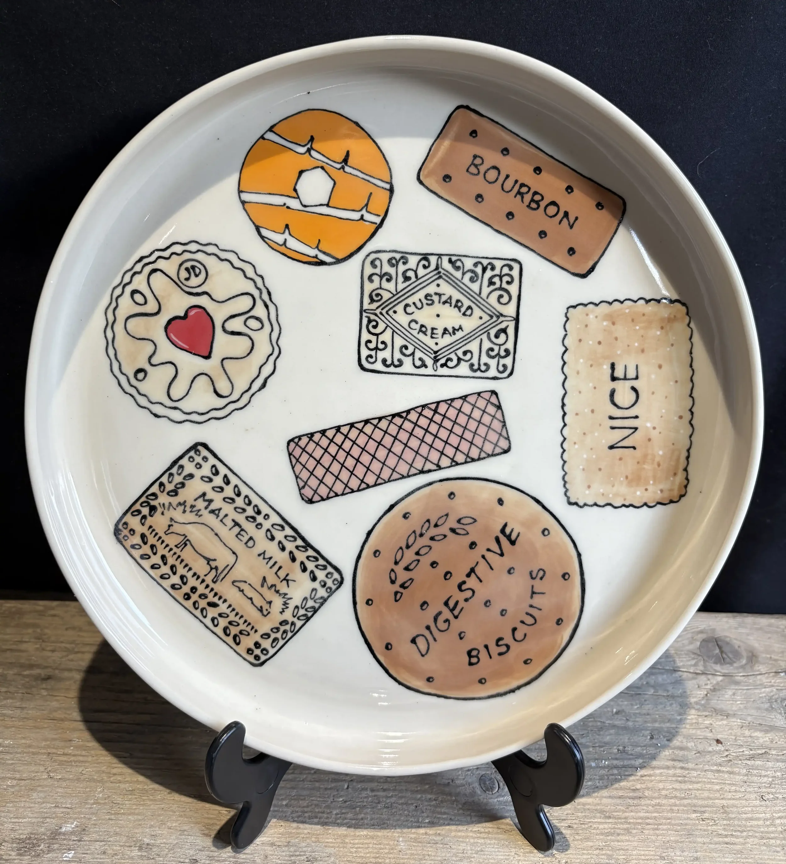 Handmade, plate with illustrations of various British biscuits labeled with their names, a Jammie Dodger, an orange Party Ring biscuit, Bourbon biscuit, Custard Cream, a NICE biscuit, a Malted Milk biscuit, Pink Wafer and a large Digestive biscuit.