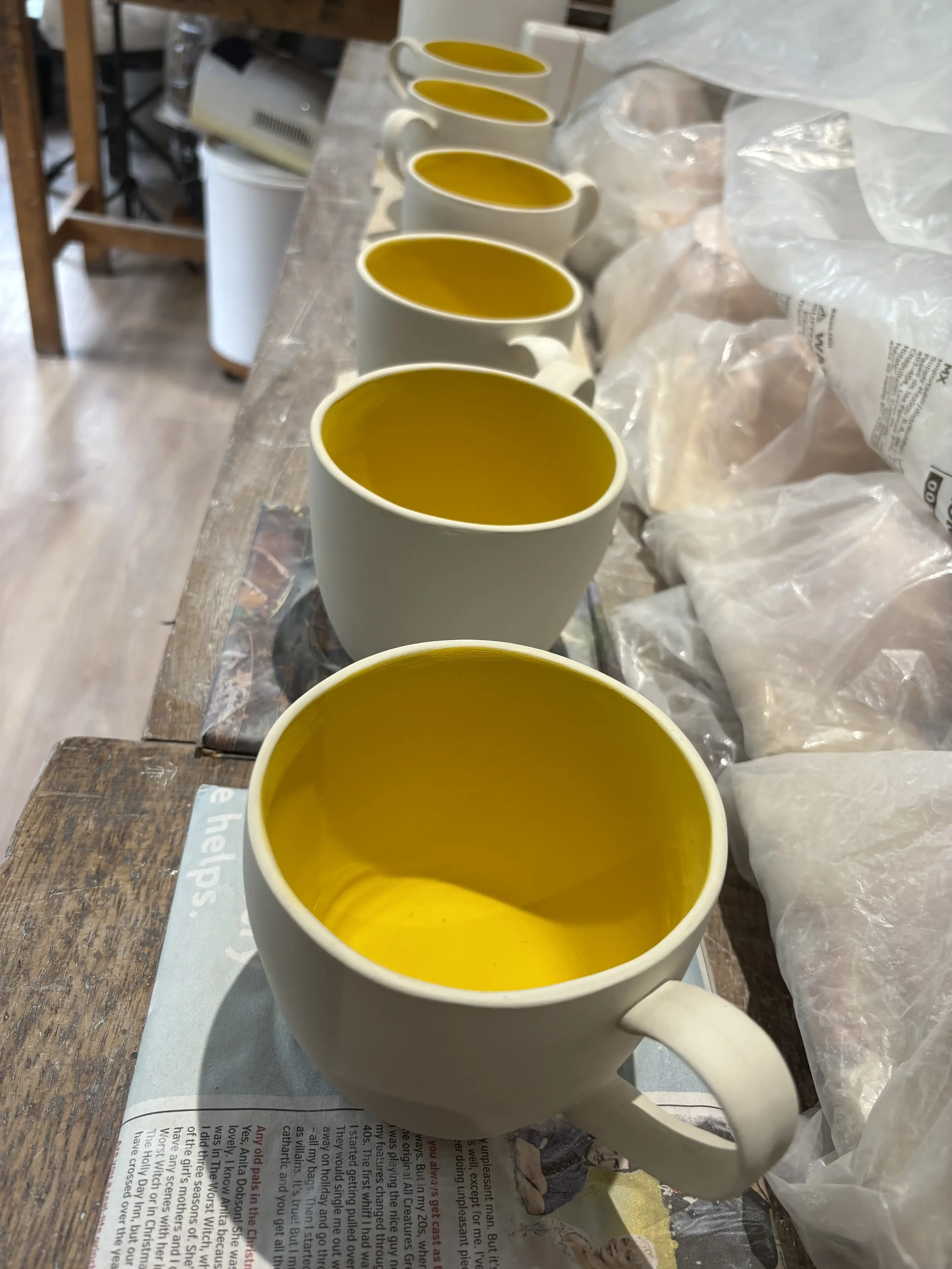 Six ceramic mugs decorated with yellow inside, lined up on a wooden surface, in a pottery studio setting.