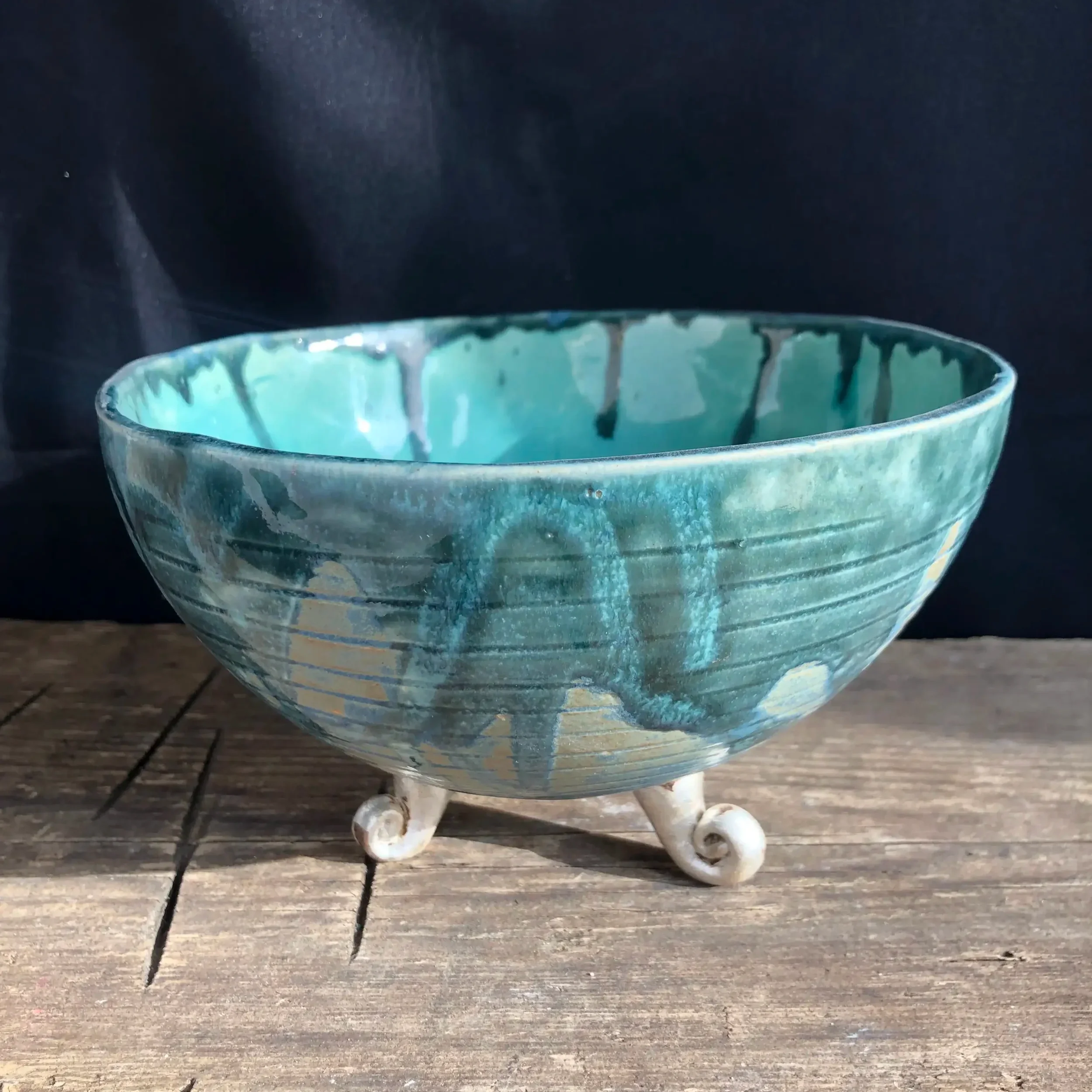 Ceramic bowl with turquoise and blue glaze on three small white curled feet, placed on a wooden surface with a dark background.