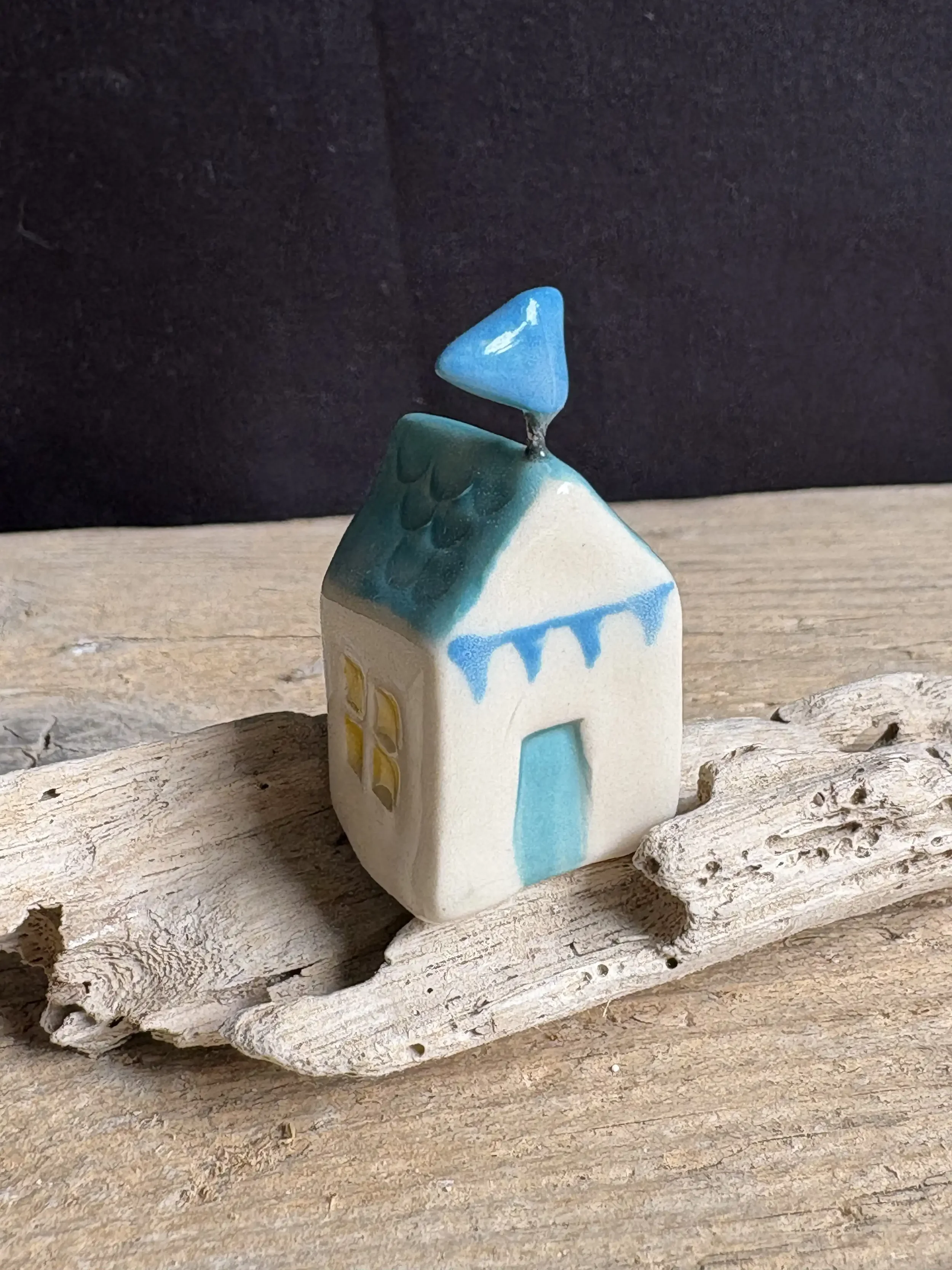 Teeny ceramic beach hut with a blue and white bunting design and small blue flag on top of the hut. It is sitting on a piece of driftwood on a wooden surface.