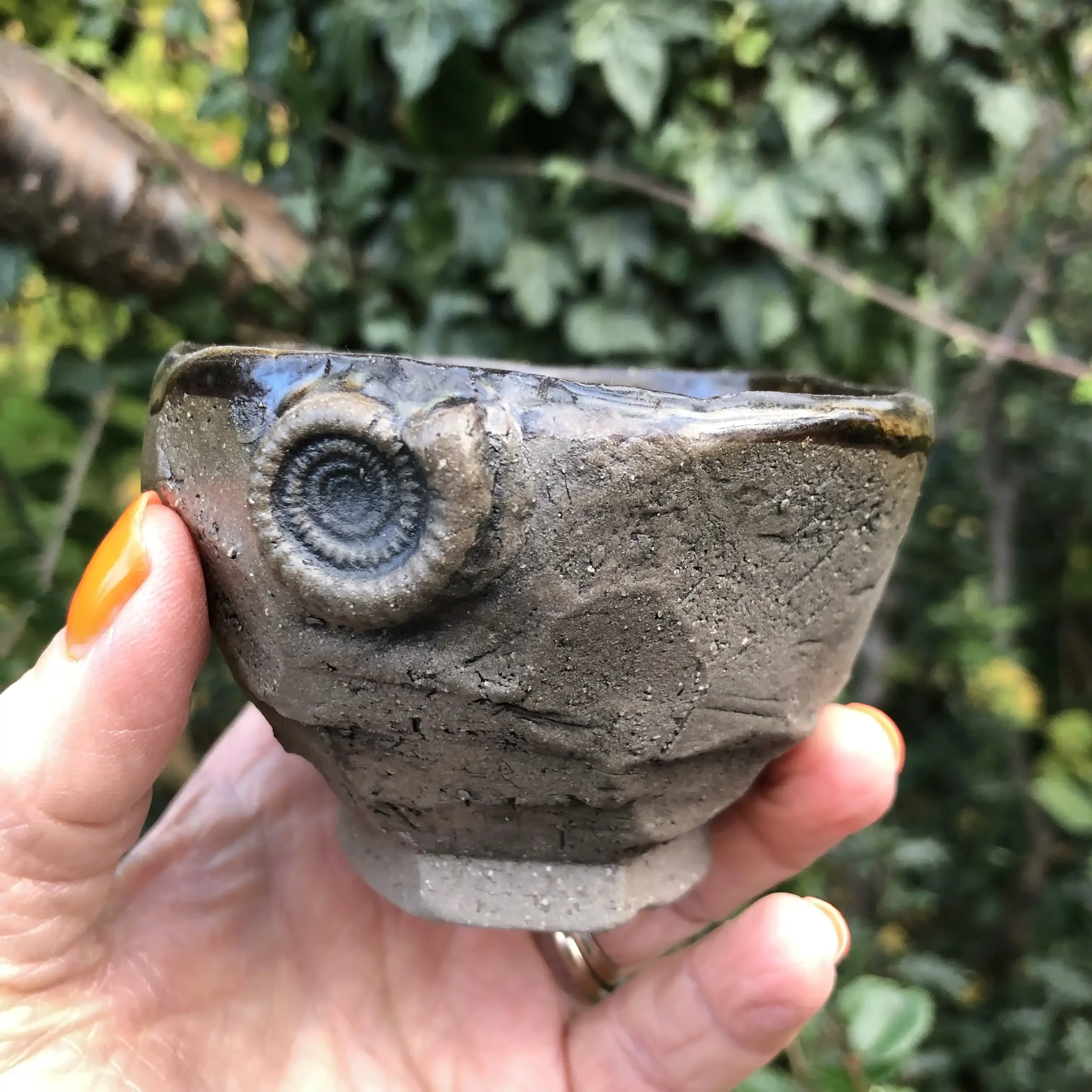 A hand holding a carved Kurinuki style cup with an ammonite on the rim, outdoors with green foliage in the background.