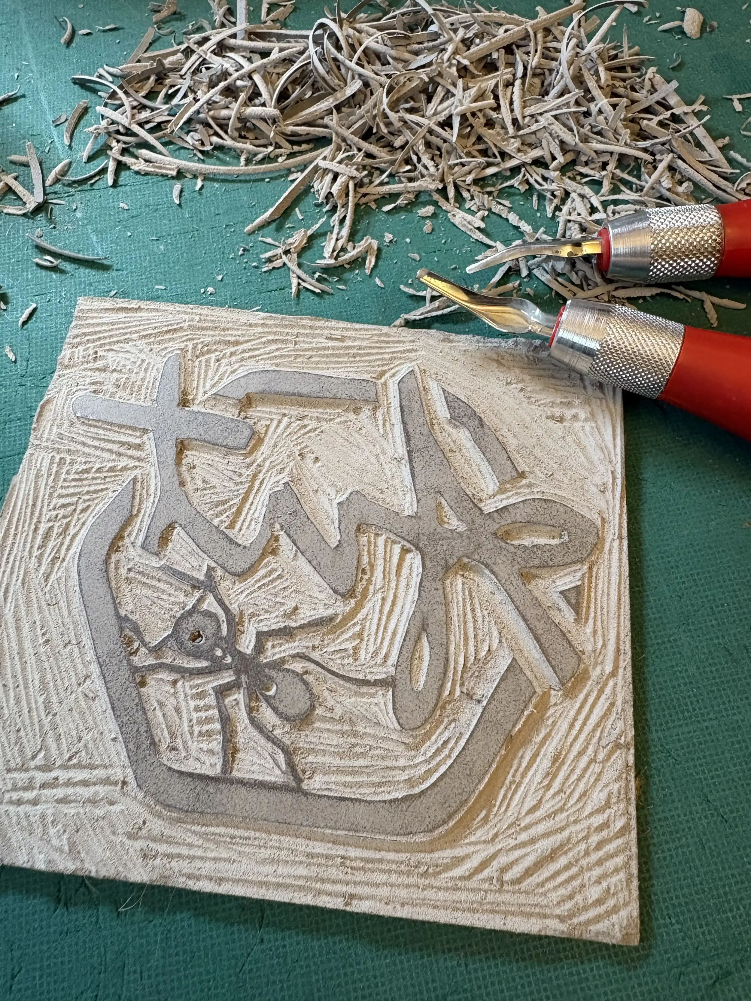 Lino cut carving of a hexagon, script 'Ant' and an ant, with carving tools and wood shavings around it.