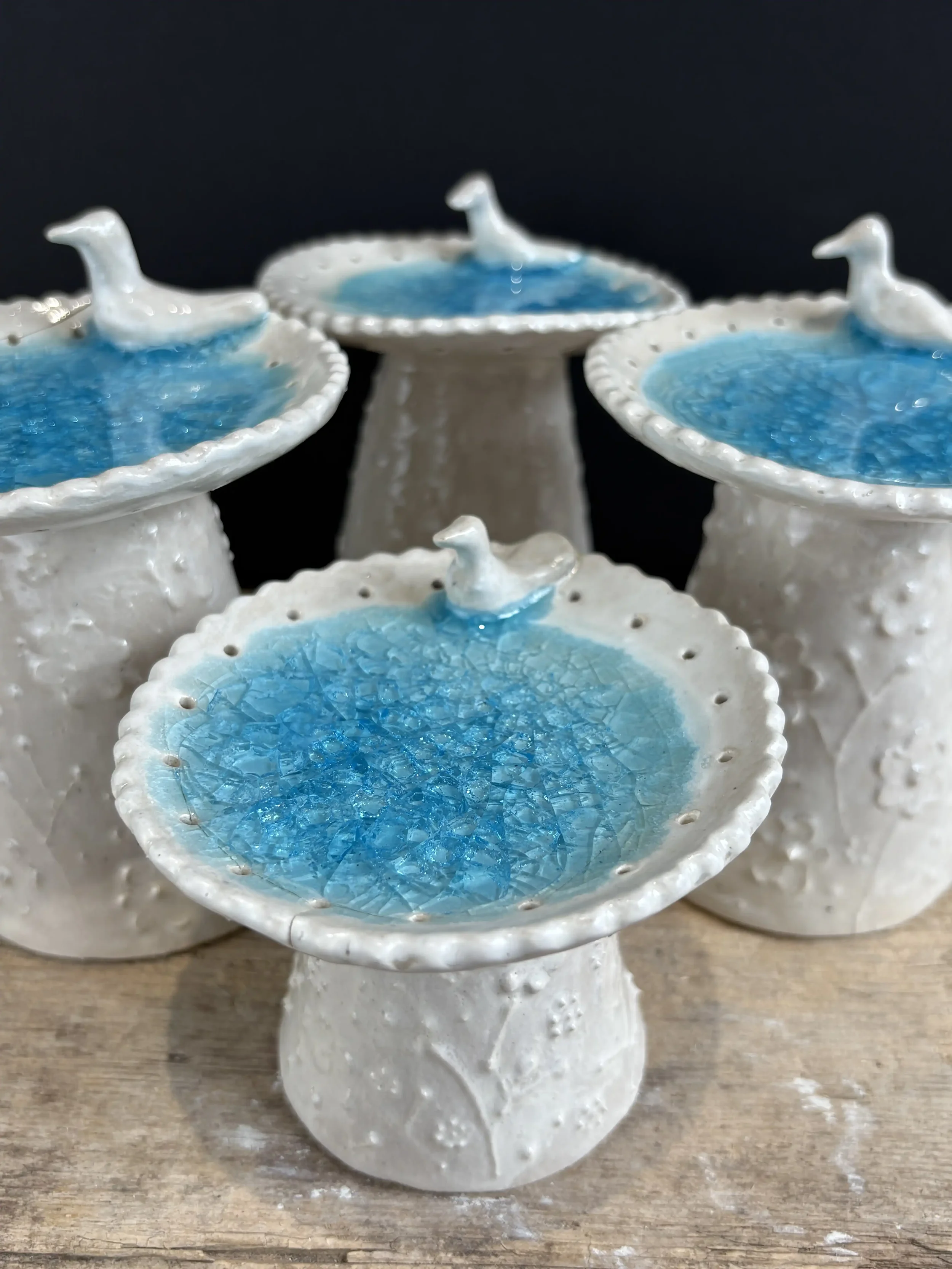 Set of four white ceramic birdbath-shaped jewellery stands with melted blue glass to resemble water inside the dish, placed on a wooden surface against a black background.
