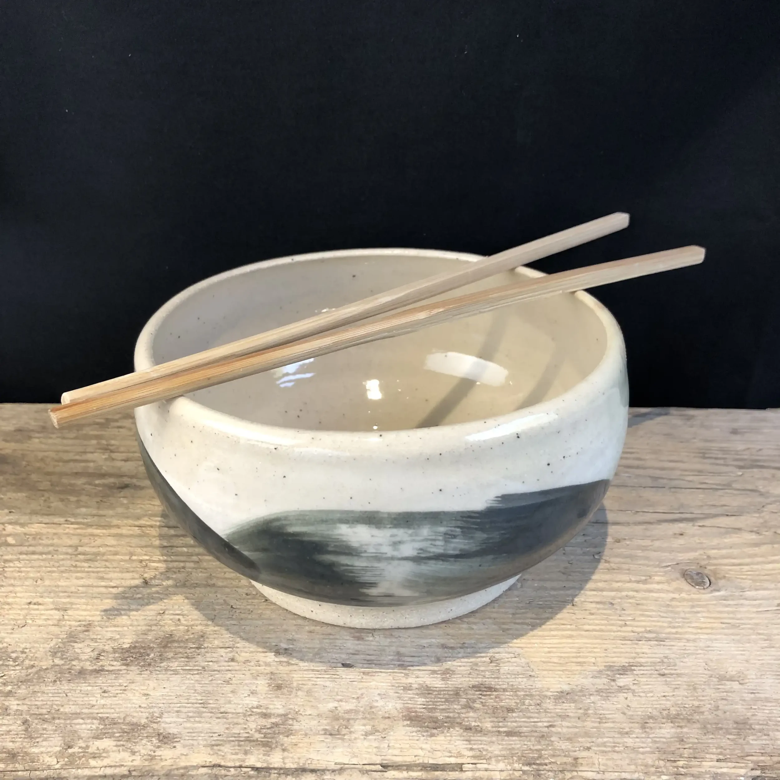 A ceramic bowl with black and white abstract brushstroke patterns. Two wooden chopsticks resting on top, placed on a wooden surface against a black background.