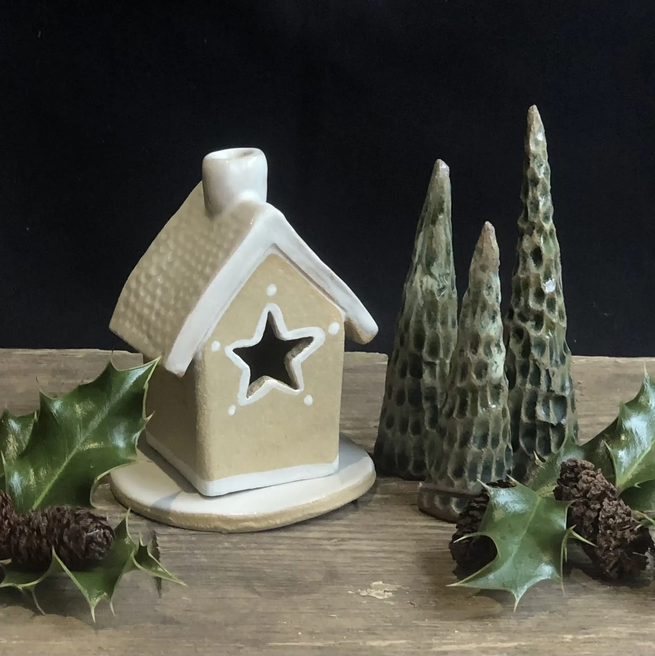 Christmas ceramic house ornament with cutout white star window, three green textured cone-shaped trees, holly leaves, and pine cones on a wooden surface against a black background.