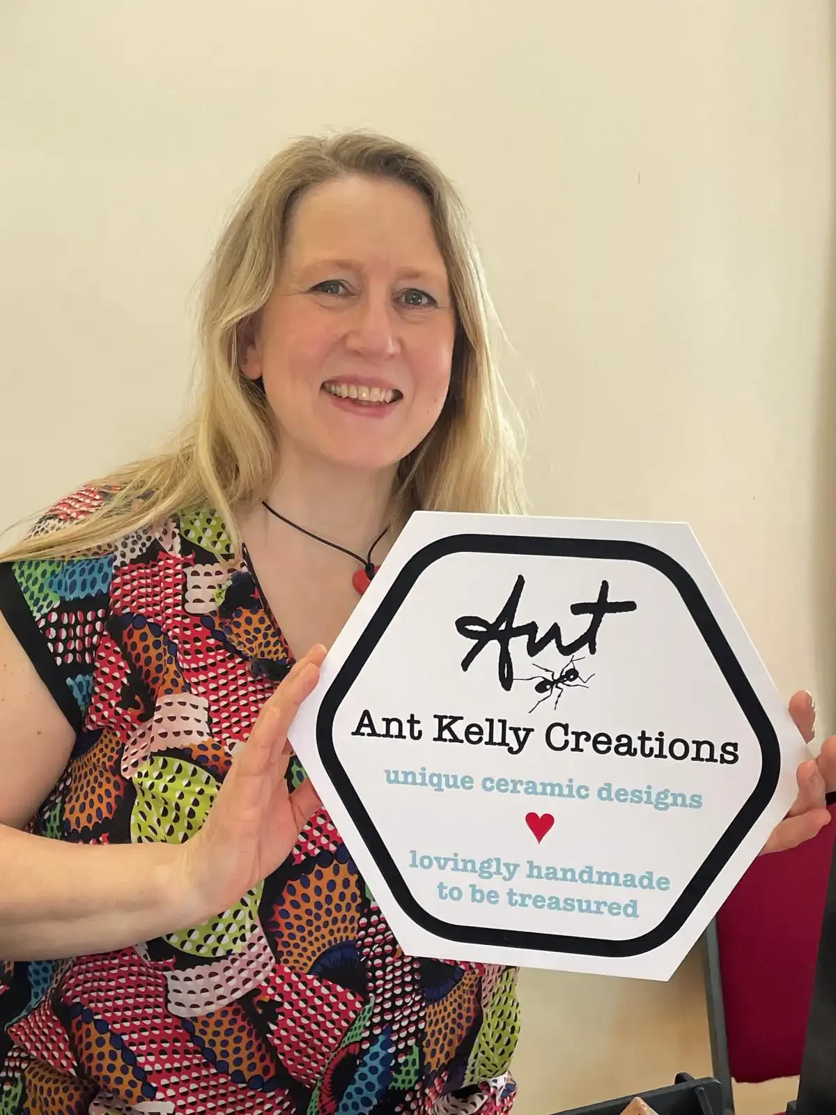 A smiling woman with long blonde hair holding a hexagonal sign that says 'Ant Kelly Creations, unique ceramic designs, lovingly handmade to be treasured,' with a small red heart and an ant illustration.