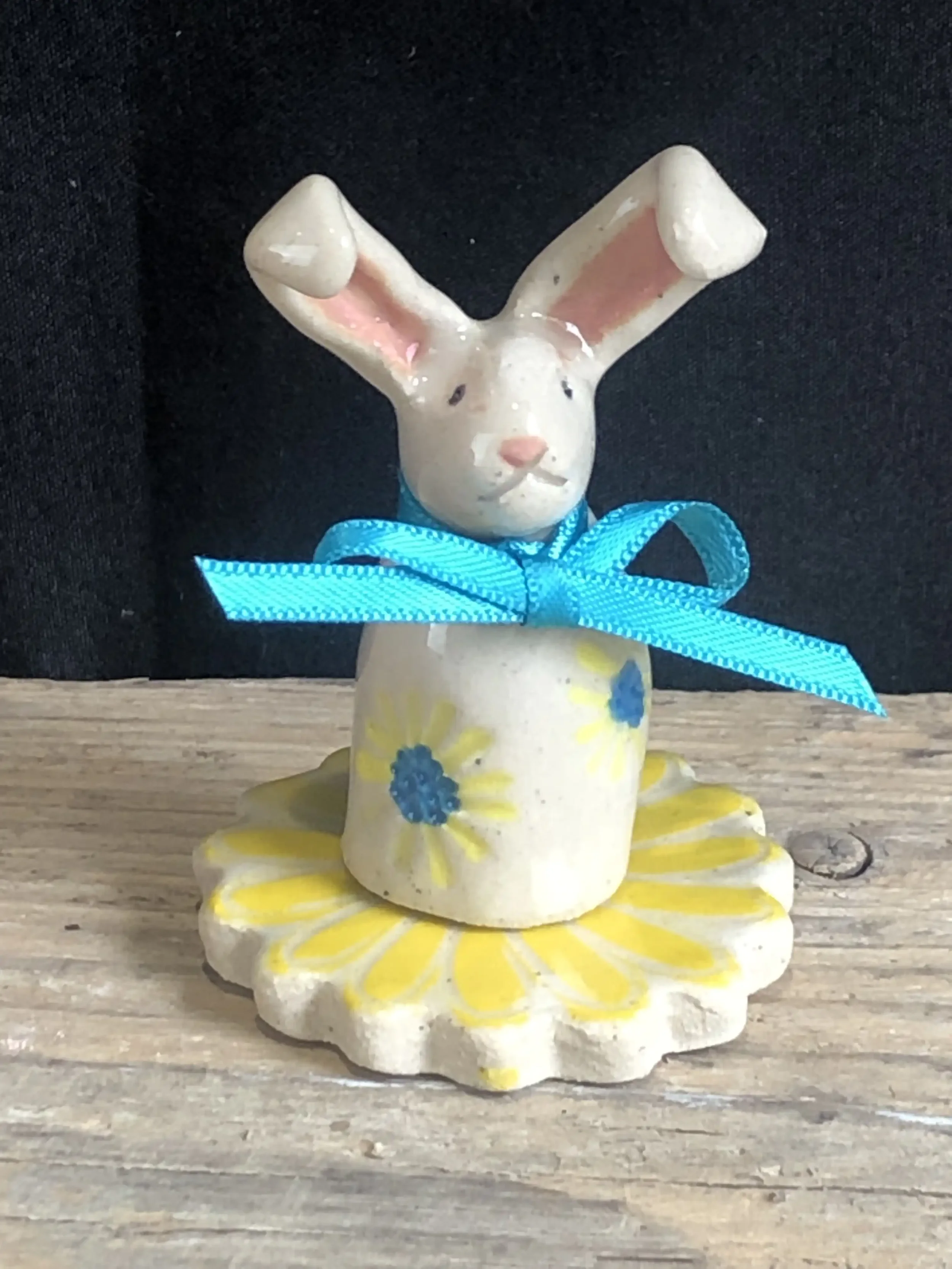 Ceramic bunny figurine with pink ears, blue bow, yellow and blue painted flowers, sitting on a wooden surface.