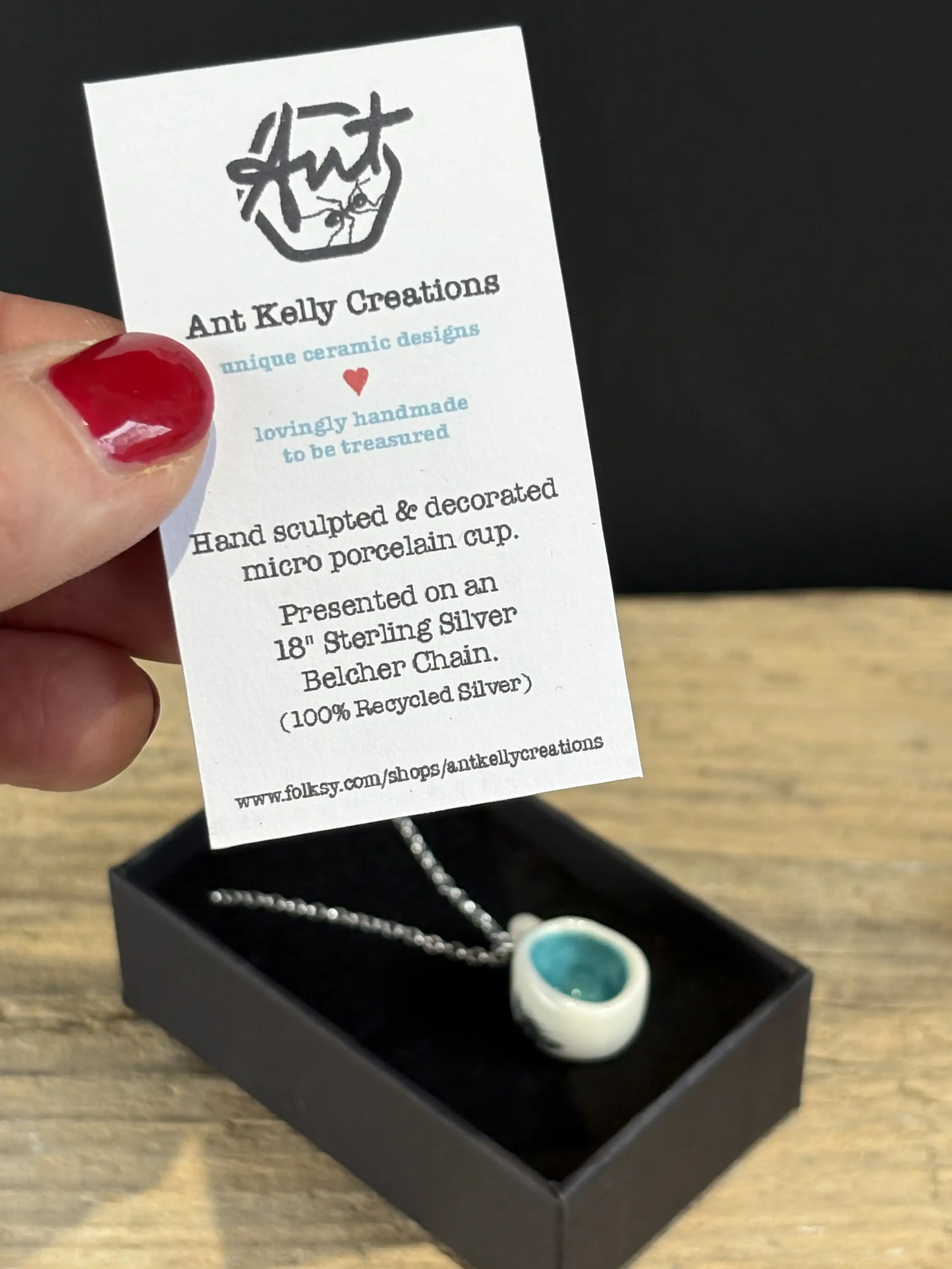 A hand holding a description card for Ant Kelly Creations porcelain jewellery. There is a small porcelain mug pendant on a chain inside a black box.