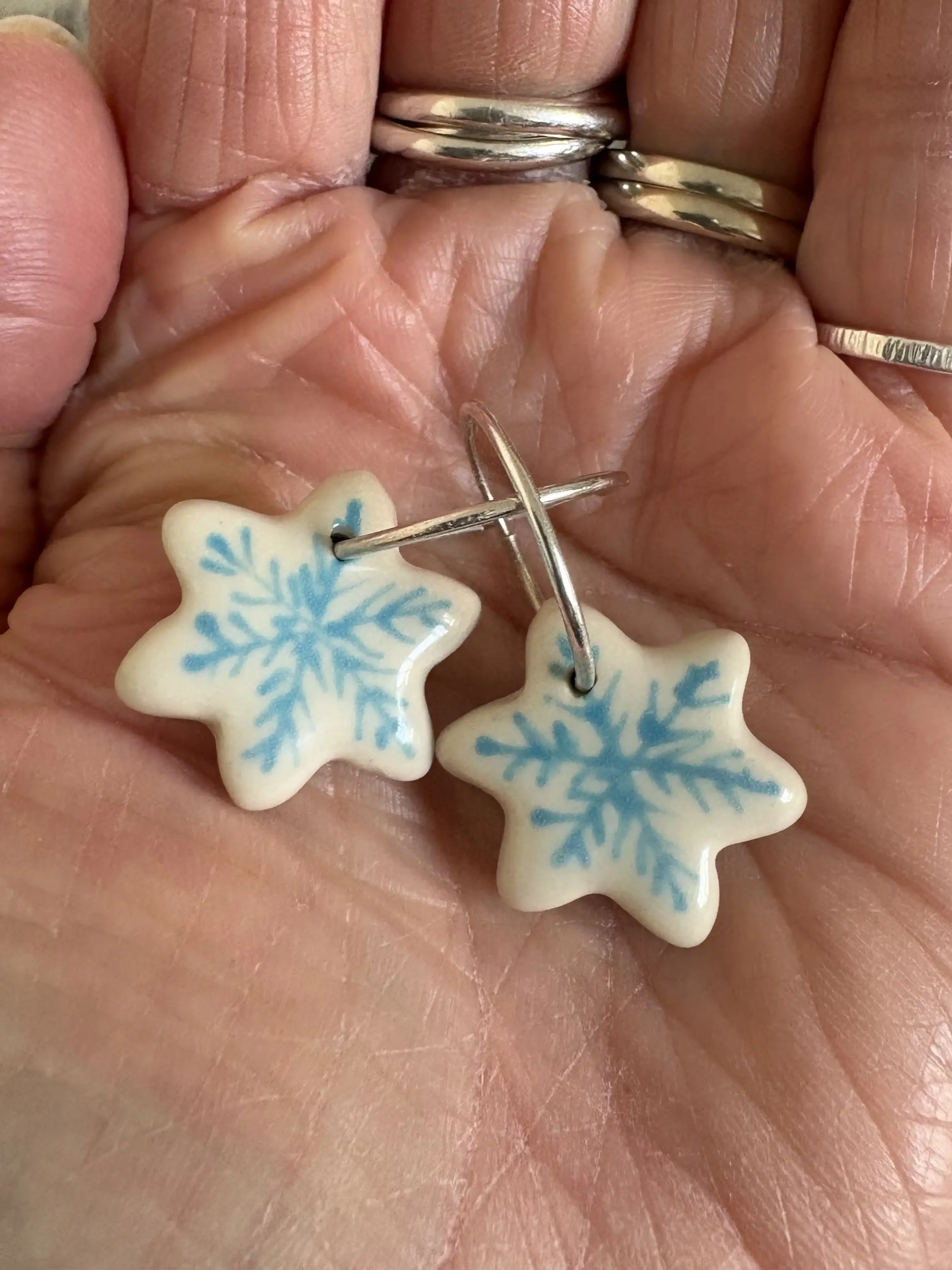 A hand holding a pair of cream-colored earrings shaped like stars with blue snowflake designs, attached to silver hoops.