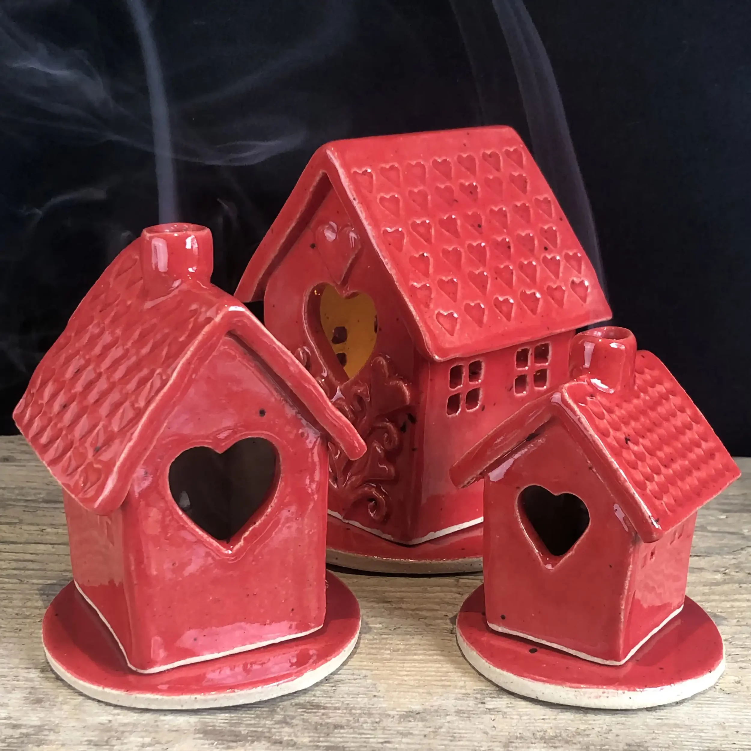 Three red ceramic house-shaped incense burners and a tea-light holder with heart-shaped cutouts, arranged on a wooden surface with a dark background.