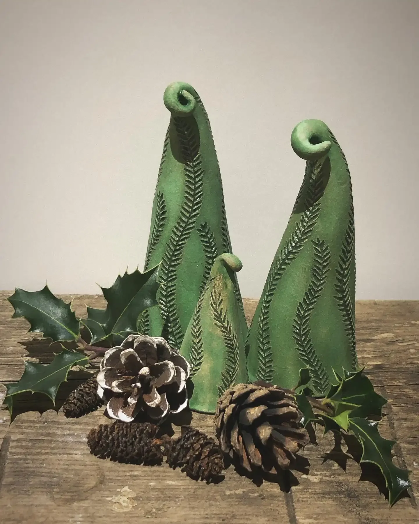 Decorative Christmas scene with three green ceramic trees with leaf patterns, surrounded by holly leaves and pinecones, on a wooden surface.