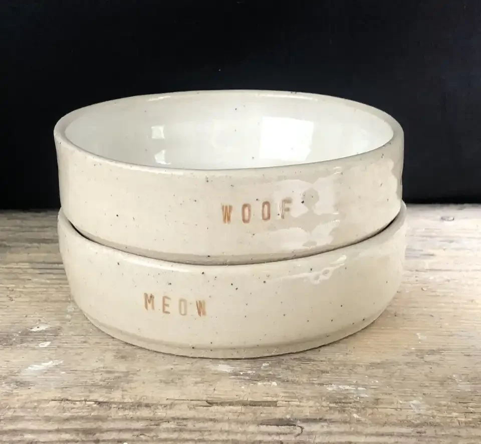 Two white ceramic pet bowls stacked on a wooden surface, labeled 'WOOF' and 'MEOW' in brown lettering.