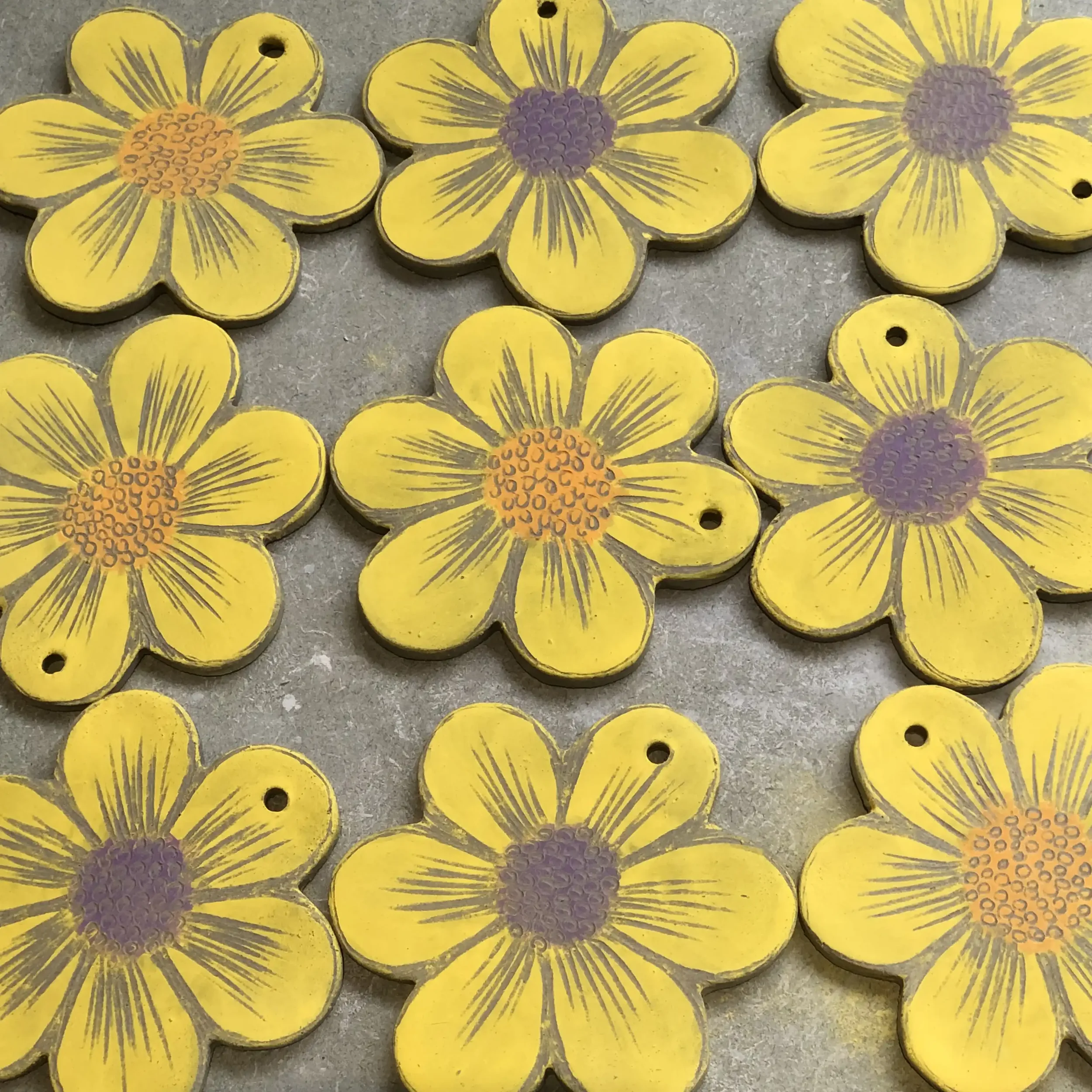 Nine decorative flower-shaped clay ornaments painted yellow with purple and orange centers, placed on a gray surface.