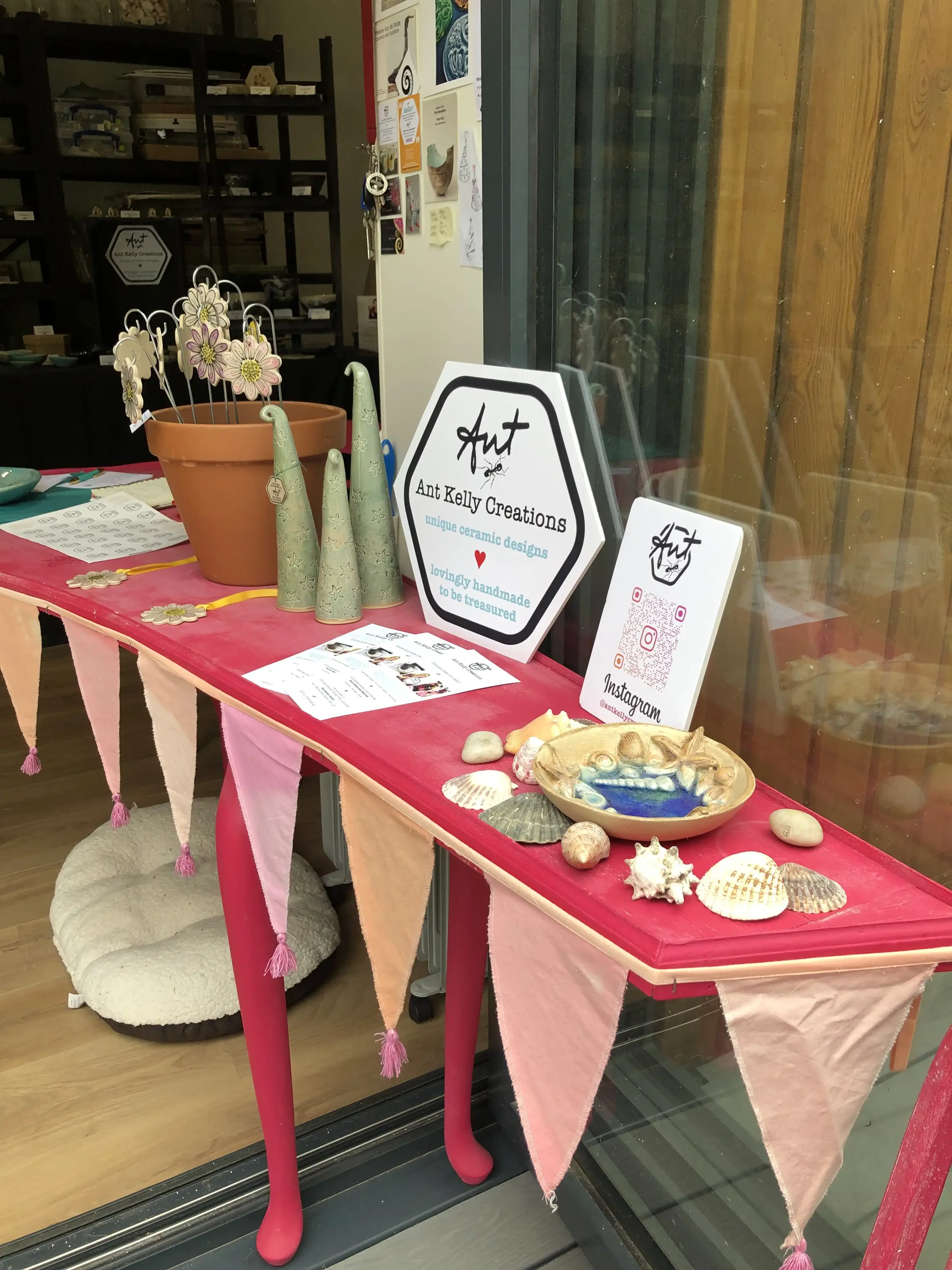 Display table with ceramic coastal-themed crafts, seashells, and signs for 'Ant Kelly Creations'.